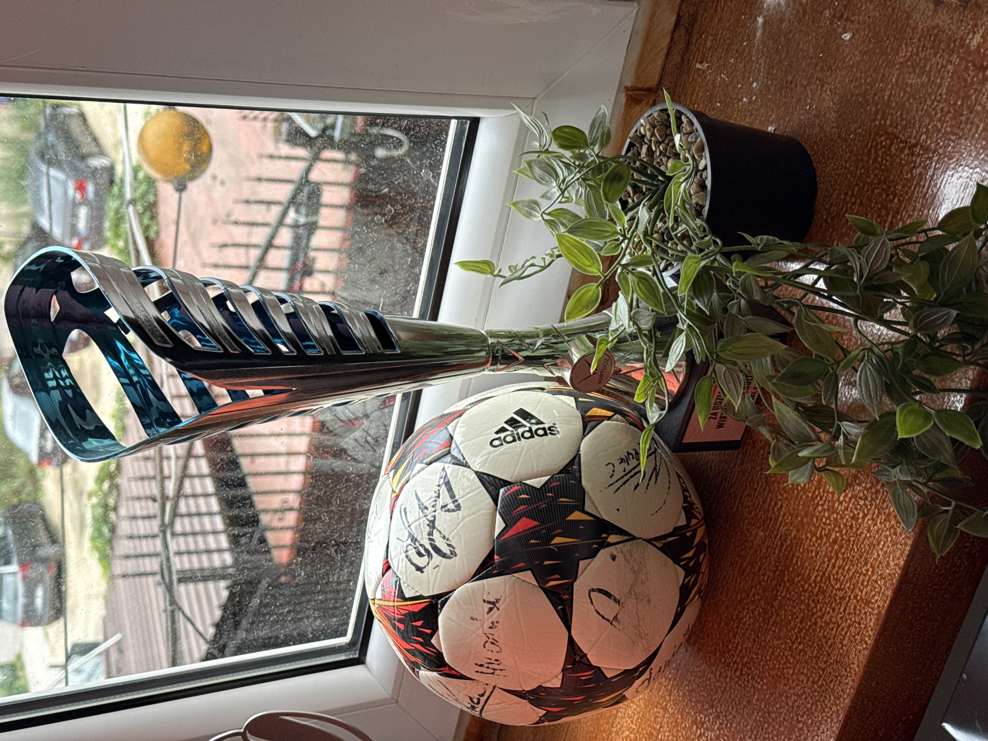 The image depicts a close-up view of a wooden windowsill. On the windowsill, there are three main objects: a signed soccer ball, a tall, decorative trophy with a metallic finish and blue accents, and a potted plant with trailing green leaves. The soccer ball, positioned to the left, has a pattern of black, red, and yellow shapes. The trophy stands to the right of the ball, appearing to be made of polished metal with interwoven blue sections. The potted plant is in a black pot filled with small stones and gravel, located to the right of the trophy. The background shows a window with a view of what appears to be an urban environment with cars and buildings visible outside, suggesting the location could be in the city of Łódź, Poland, as indicated by the inscription on a small plaque below the trophy which reads "ZA UCZCIWĄ GRĘ WIRTUO..." which translates to "FOR FAIR PLAY VIRTUO...". The window panes are visibly dirty, with water droplets present, indicating it might be raining or has recently rained. The overall lighting suggests an indoor setting near a window.