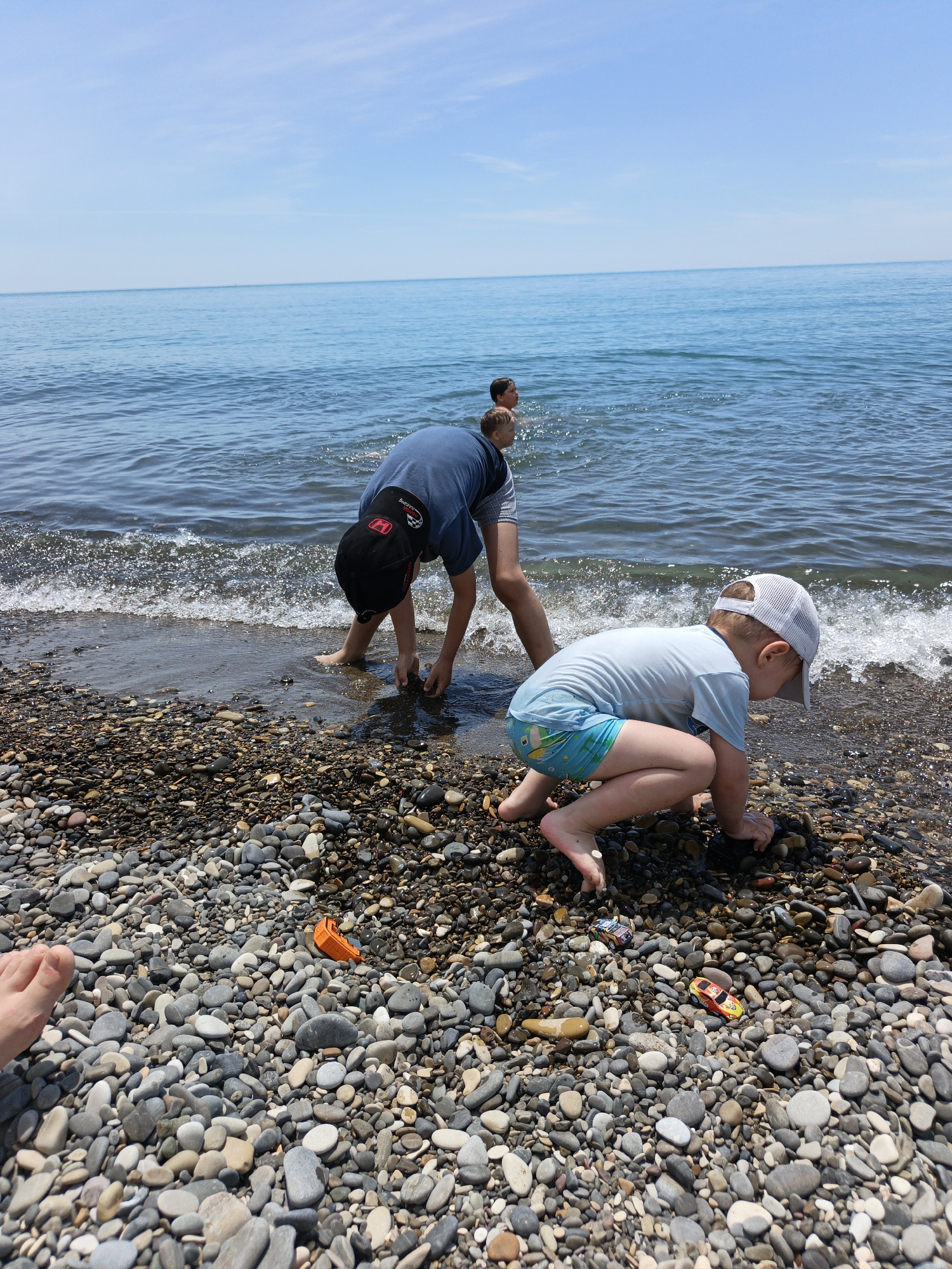 The image depicts a sunny pebble beach scene in Krinitsa, Russia. In the immediate foreground, a bare human foot rests on light-colored, smooth pebbles. To the right, a young child, wearing a light blue short-sleeved shirt, blue and green patterned swim trunks, and a white mesh baseball cap, crouches barefoot at the water's edge, intently focused on the wet pebbles. Two small toy cars (one orange, one blue) and an orange boat-like toy are visible on the pebbles near this child. Slightly behind and to the left, an older child or pre-teen, wearing a dark blue short-sleeved shirt and a black baseball cap with a red "H" logo, bends forward with hands submerged in the shallow breaking waves, appearing to collect or examine items. Further into the calm, clear blue sea, two additional individuals are partially visible, with heads and shoulders above the water. The horizon line is clearly defined, separating the serene sea from a light blue sky with faint, wispy clouds. Small, white-crested waves break gently onto the pebble-covered shore.