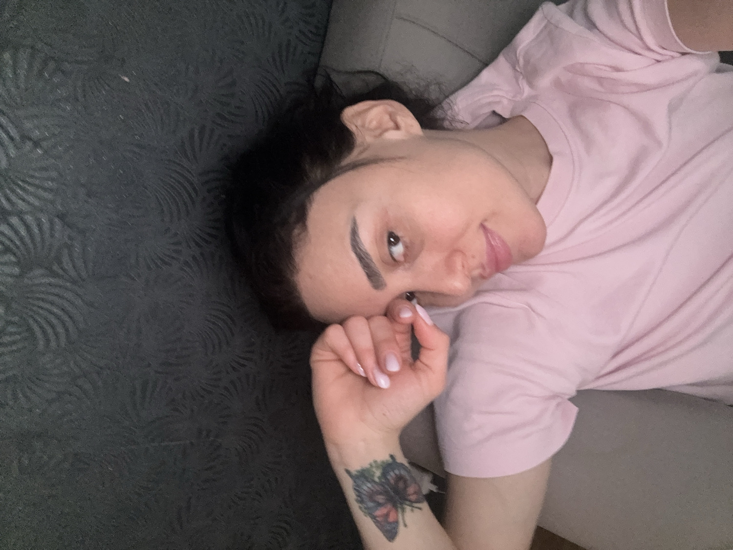 An intimate, high-angle portrait features a young woman lying down in an indoor setting, looking directly at the viewer with a relaxed and engaging expression. Her head is tilted, and she has dark, wavy hair, and prominent, dark eyebrows. She wears a simple, light pink short-sleeved t-shirt. Her left hand is gently raised, with light-colored polished nails, and on her left forearm, a colorful tattoo of a butterfly is clearly visible, featuring shades of purple, green, and red. She has a soft smile or smirk on her lips, and her gaze is direct and inviting.
The background is a dark grey or black textured surface, possibly a sofa or bed, with an intricate, swirling pattern resembling stylized fronds or abstract designs. A lighter grey surface is also visible to her right, contributing to the indoor, domestic atmosphere. The lighting is even, suggesting an interior space without harsh direct light. No specific location cues are visible within the image, but it is provided that the context is Tbilisi, Georgia. There is no visible text.