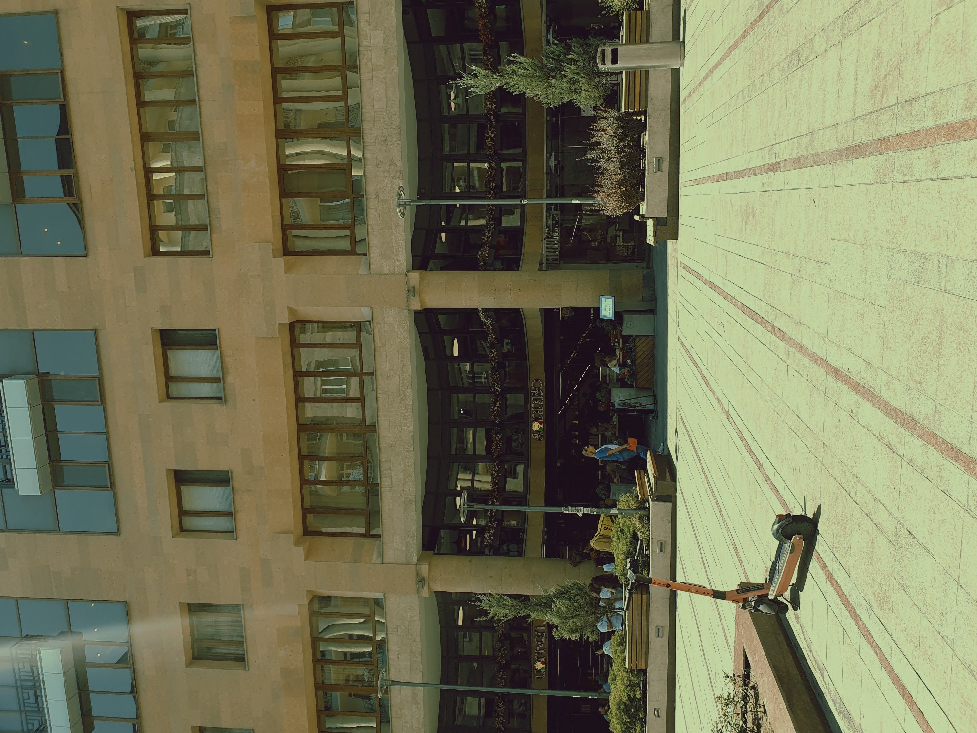 In a city square in Yerevan, Armenia, an orange electric scooter rests on a light-colored tiled walkway marked with red stripes. Behind the scooter, a modern building with a beige stone facade features numerous windows. The ground floor of the building has a dark glass facade, suggesting shops or cafes, with people seated at tables visible inside. Several trees and benches are placed along the building's facade, and a few individuals are walking or sitting in the area. The overall scene depicts a sunny urban environment.