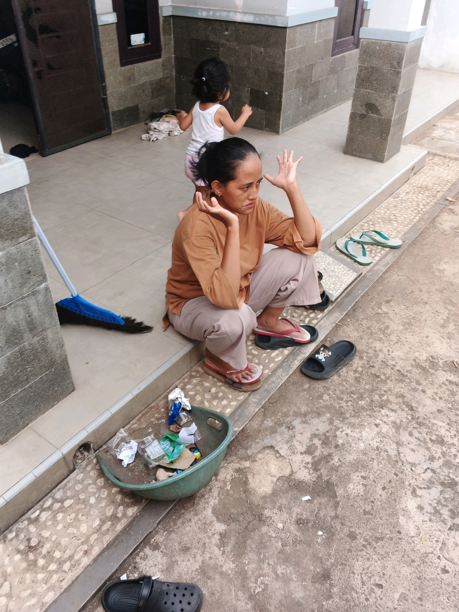 In Kadu, Indonesia, a woman is sitting on the ground in front of a building, with a child playing in the background. The woman is wearing a brown shirt and light-colored pants, and she is looking to the side with her hand raised. The child is wearing a white tank top and pink shorts, and is facing away from the camera.
The scene appears to be an outdoor area with a tiled floor and a concrete walkway. There is a broom leaning against a wall, and a green plastic basket filled with trash on the ground. Several pairs of flip-flops are scattered around the area. The building in the background has a dark door and stone walls. The overall environment suggests a residential or public outdoor space.