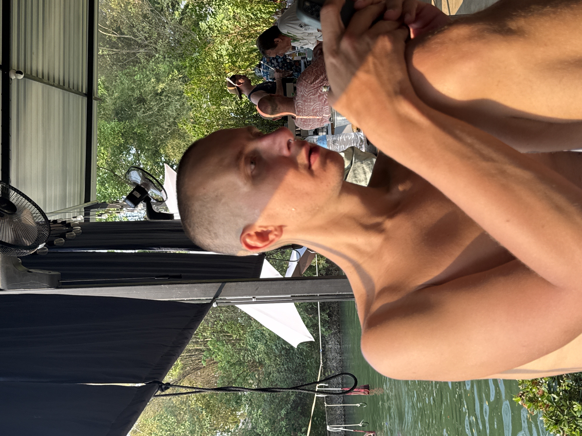 The image shows a shirtless man with a shaved head getting a haircut outdoors. The location appears to be a covered patio or deck with a view of a body of water and trees. Other people are present in the background at tables. Electric fans are visible on the deck. Cables and possibly a dock are visible along the water's edge. The image was taken during daylight. This location is in the city of Tambon Nam Phrae, Thailand.