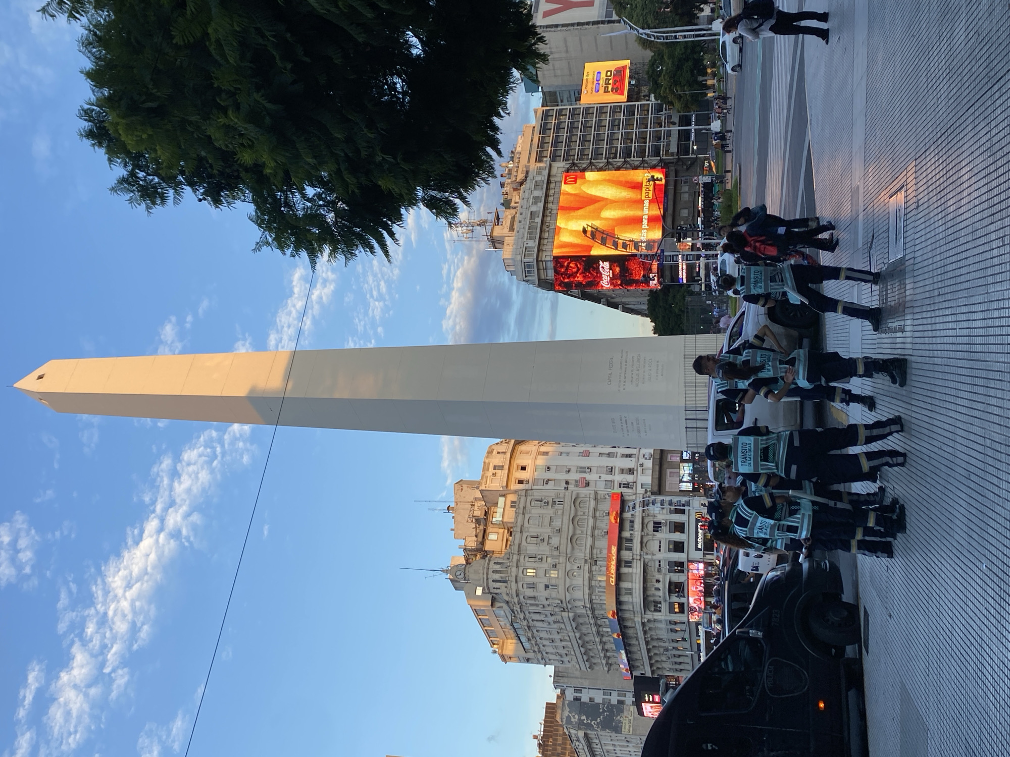 The image shows the Obelisco de Buenos Aires, a historical monument in the city of Buenos Aires, Argentina. The monument is a tall, white, rectangular obelisk set against a blue sky with scattered clouds. In the background, there are buildings with digital billboards. In the foreground, there is a group of officers wearing vests that read "TRANSITO DE LA CIUDAD." A black vehicle with "POLICIA" on the side is partially visible at the bottom.