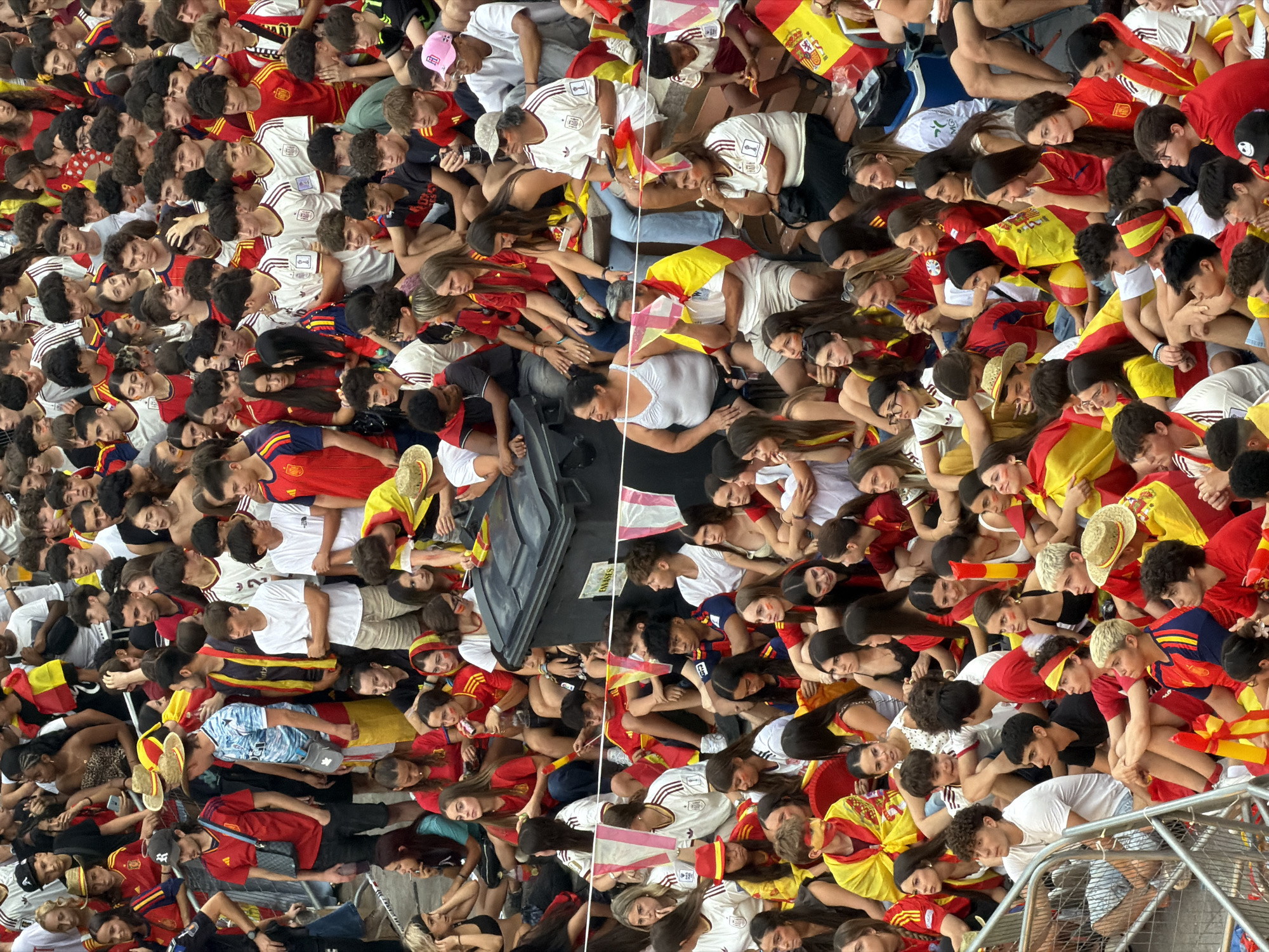 The image depicts a large crowd of people, predominantly young adults and teenagers, gathered outdoors. Many are wearing red and yellow, the colors of the Spanish flag, suggesting a sporting event or national celebration. Some individuals have face paint in these colors. The crowd is densely packed, with people sitting and standing. Several are holding Spanish flags or wearing them as capes. In the background, there is a large black trash bin and some fencing. The lighting suggests it is daytime. The context implies a public event in Las Rozas de Madrid, Spain, likely a football match or a similar patriotic gathering.