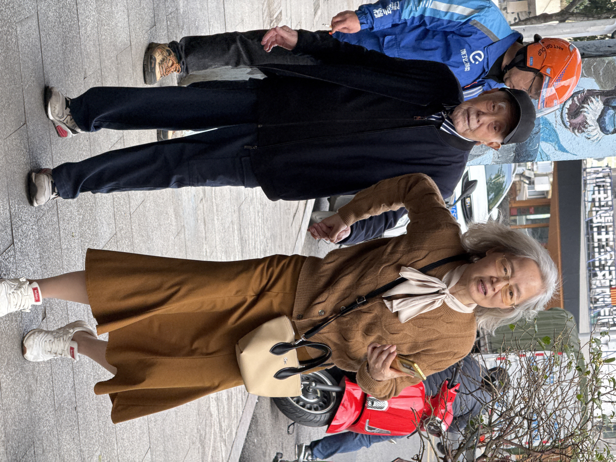 An elderly man and a middle-aged woman are walking on a paved sidewalk in Jiulongpo District, China. The man, positioned on the left, wears a black baseball cap, a dark blue zip-up jacket over a black and white striped shirt, dark blue cargo pants, and grey athletic shoes. His right hand is clasped by the woman's left hand. The woman, to his right, has grey hair, wears gold-rimmed glasses, a brown cable-knit cardigan, a light beige neck scarf tied in a bow, a brown midi skirt, and white New Balance athletic shoes. She carries a small beige Lacoste handbag with black handles over her left shoulder and holds a gold-colored smartphone in her right hand. She is looking forward, slightly towards the viewer, with her mouth slightly open. To the left and slightly behind the man, a delivery driver in a blue uniform with Chinese characters ("饿了么") and an orange helmet with "ANT GRUP" text is visible, wearing dark pants and tan hiking-style shoes. The immediate background features a wall with a partially visible blue and white mural. Further back on the right, a red scooter is parked or in motion, partially obscured by a tree with sparse branches. Other vehicles, buildings, and overhead utility lines are visible in the distance under daylight conditions.