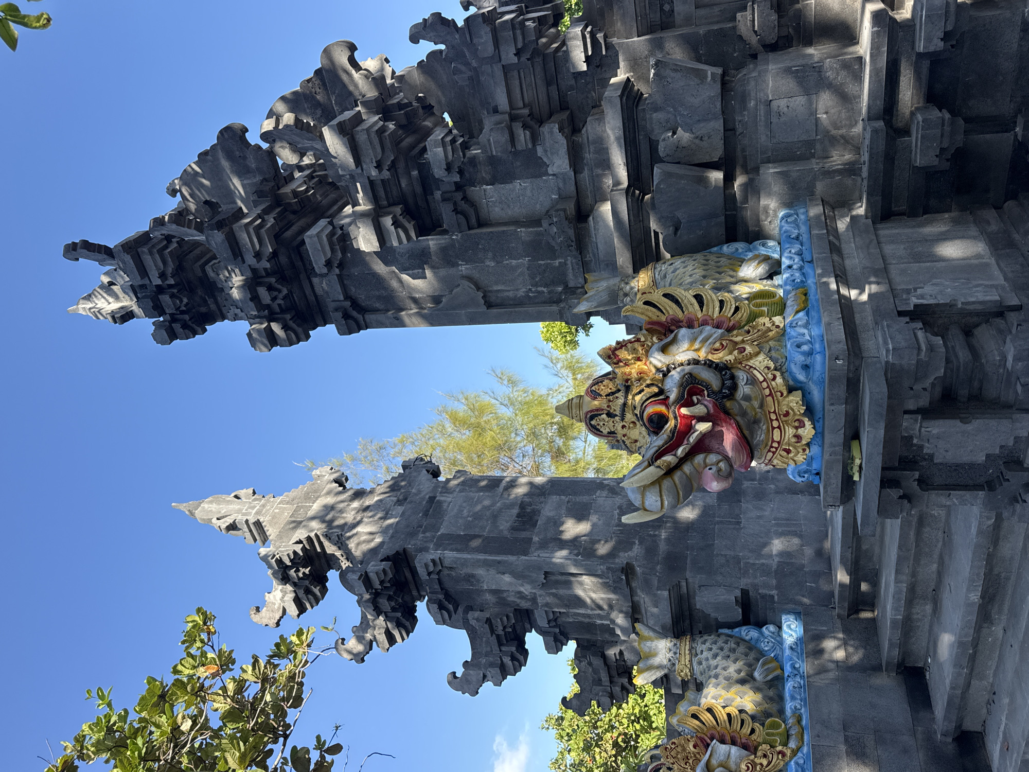 The image captures a grand traditional Balinese temple gate, known as a candi bentar, against a clear blue sky. The gate is constructed from dark gray, intricately carved stone, featuring a tiered, stacked design with multiple receding layers that create a sense of depth and majesty. Its surfaces display textured details and sharp angles, characteristic of Balinese architecture.
Flanking the entrance, two vibrant, multi-colored guardian statues are visible. The more prominent one on the right depicts a mythical creature with a fierce expression, featuring large, red-rimmed eyes, an open mouth revealing a red tongue, and elaborate gold, red, and white facial details. Its body is adorned with blue and gold scales or patterns. A similar, partially visible statue is positioned on the left side of the gate.
The scene is bathed in bright daylight, casting distinct shadows across the stone structure, indicating clear weather. Green foliage from trees is visible around the gate, particularly in the foreground and background against the blue sky. No people or activities are present, and there is no visible text. The architectural style and guardian figures are strong cultural indicators for Kuta Selatan, Indonesia.