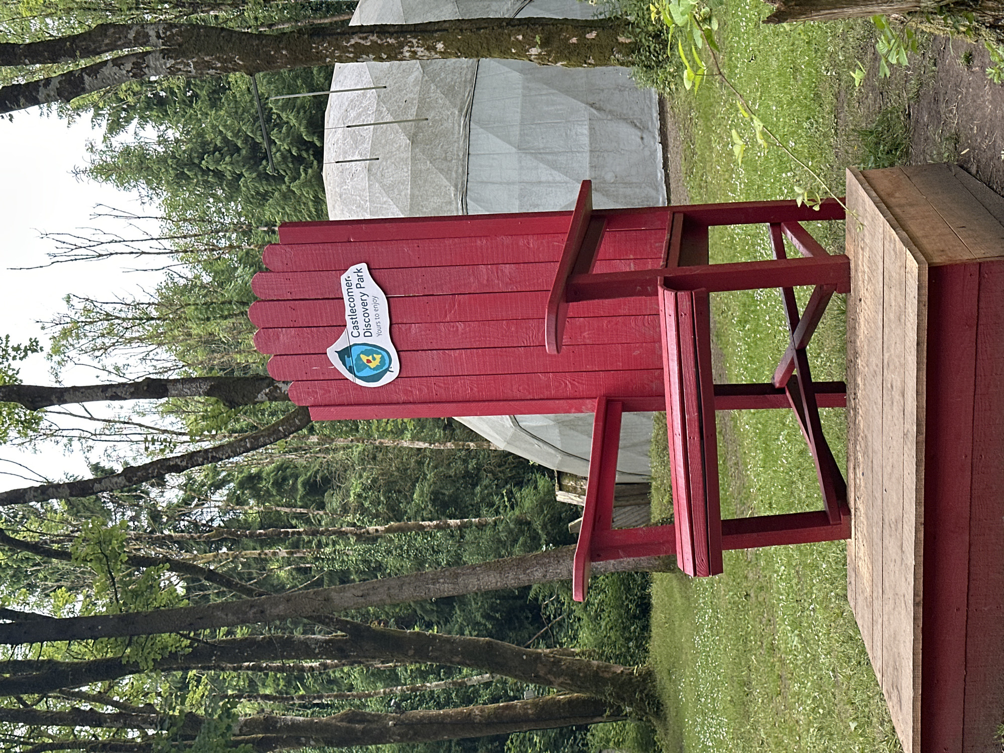An oversized red wooden Adirondack chair stands on a wooden platform in front of a geodesic dome structure. A sticker on the back of the chair reads "Castlecomer Discovery Park Yours to enjoy." The chair is set in a grassy area with trees and foliage in the background. The scene appears to be in a park or recreational area in Castlecomer, Ireland.