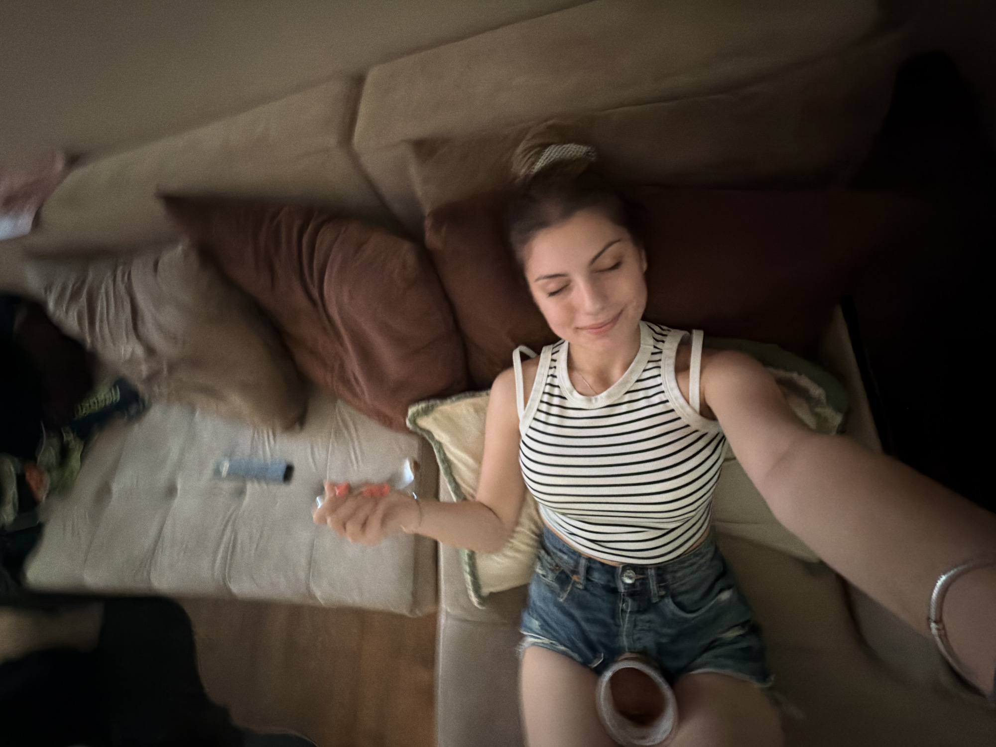 The scene depicts an indoor environment in the city of Tsarevo, Bulgaria, with a single adult female lying on a multi-sectional sofa. The woman, with a light complexion and brown hair secured with a light-colored scrunchie, is positioned on her back or slightly to her right side, eyes closed, and a subtle smile on her face. She is wearing a white tank top with thin black horizontal stripes and blue denim shorts. Her right arm is extended towards the viewer, indicating she is capturing a selfie. Her left hand, with fingernails painted an orange/coral hue, holds a silver fork. Resting on the sofa near her left hip is a light-colored cup, possibly containing a dark brown liquid. On a light beige section of the sofa, behind her left arm, a light blue/grey cylindrical object, resembling a vape pen, is visible. The sofa features varying upholstery shades, including light beige tufted cushions and darker brown/taupe sections, and is adorned with several throw pillows of different brown and cream tones, one with a green-trimmed border supporting her left arm. Blurred objects, including a blue packaged item, are scattered on the further sections of the sofa. The floor visible in the upper left is light-toned, possibly wood, and the walls are a plain, light color.