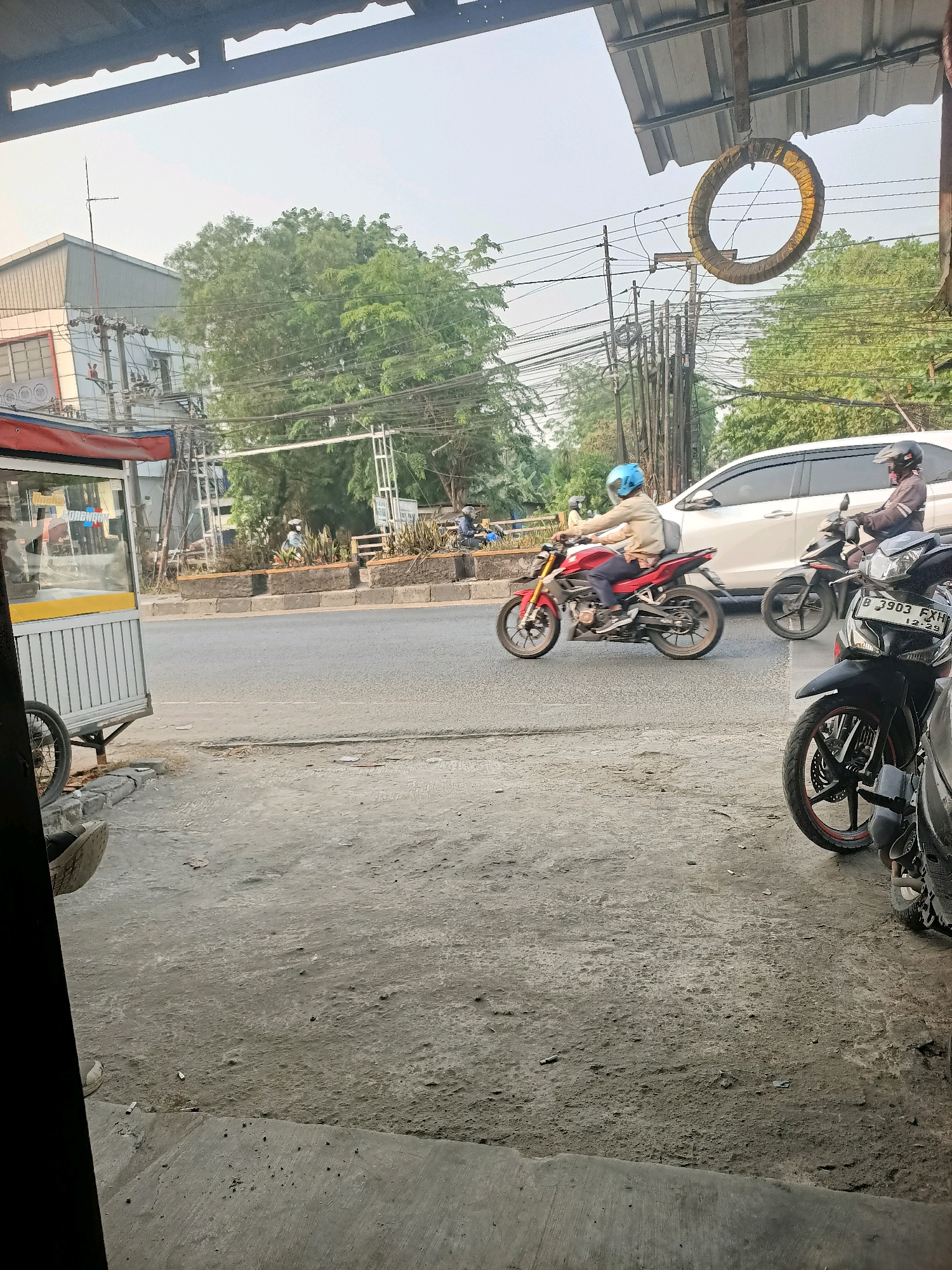 The image shows a street scene in Gandasari, Indonesia, with a focus on the road and a sidewalk area. In the foreground, a part of a motorcycle with license plate "B 3903 FXH" is visible on the right. Further down the road, a person wearing a blue helmet is riding a red motorcycle. To the left, a white car is driving. Behind the road, there are buildings, trees, and numerous electrical wires. A street vendor cart is positioned on the left side of the frame. The ground is paved with concrete and dirt. The overall atmosphere suggests a typical day in an urban or semi-urban environment in Indonesia.