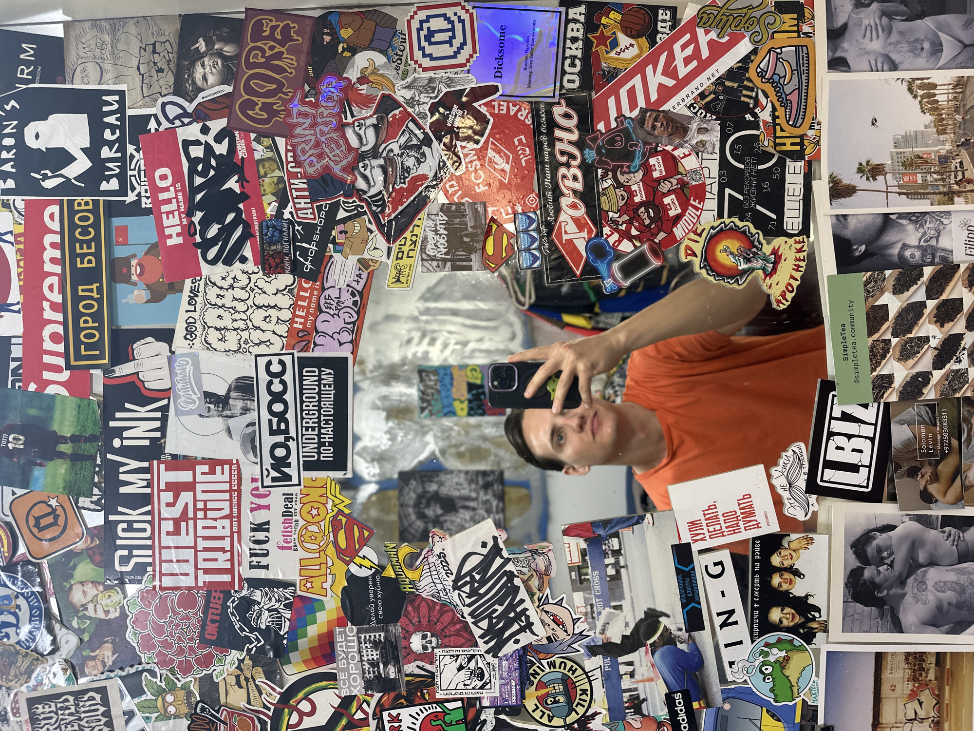 A person with short dark hair, wearing an orange t-shirt, is taking a selfie in front of a mirror covered in stickers and images. The stickers and images include various brands, logos, graffiti-style art, and photographs. Some brands featured on the stickers are "Supreme," "Adidas," "Hello," and "Dickson." The surrounding environment suggests an urban setting, with a partial view of what looks like Tel Aviv. The images include photographs of couples, palm trees, and various objects.