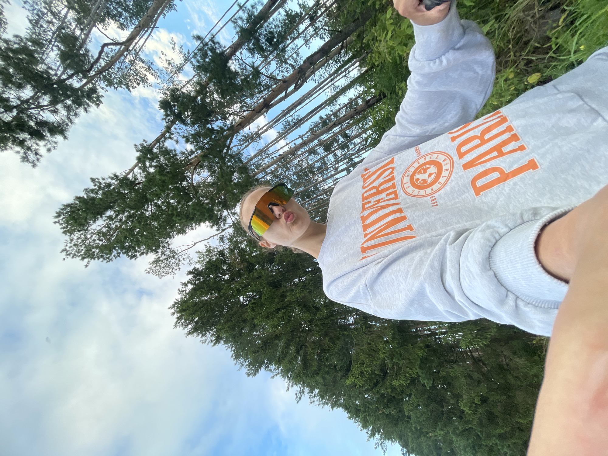 A person is taking a selfie in a forest, holding a phone with their left hand which is out of frame. The person is wearing a light grey sweatshirt with orange lettering that says "UNIVERSITY" and "PARIS". They are also wearing reflective, oversized sunglasses. The background consists of tall, thin pine trees reaching towards a cloudy blue sky. The location is the outskirts of Podshivalovo, Russia.