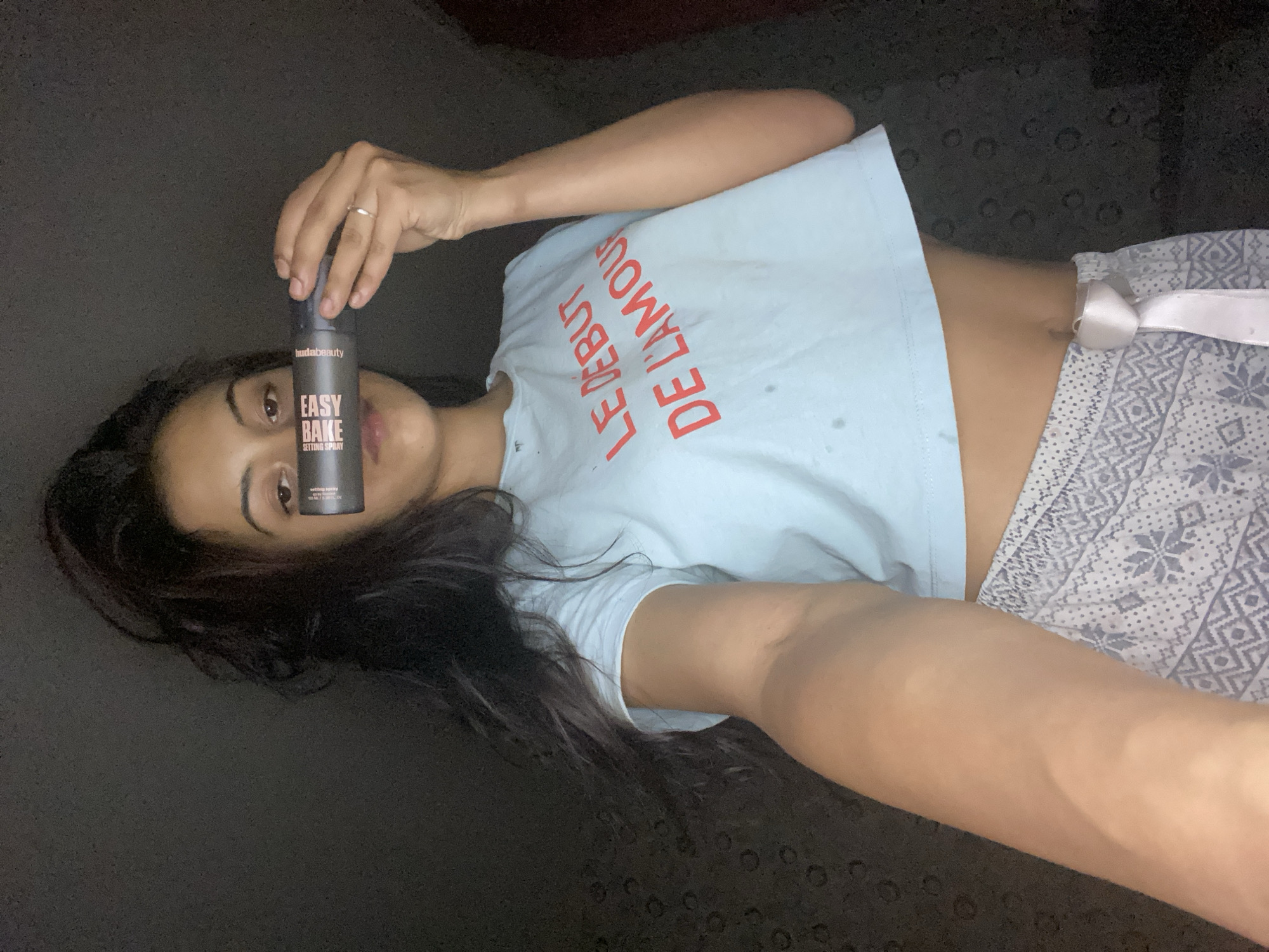 A medium shot captures a person lying down, holding a cosmetic product bottle upwards. The individual is a woman with long, dark hair spread out, looking directly at the camera with a neutral to slight smile. She wears a light blue cropped t-shirt with red text that reads "LE DEBUT DE L'AMOUR" (French for "The beginning of love") and patterned pajama bottoms with a white ribbon tie. A silver ring is visible on her right hand, which holds the product bottle.
The bottle is dark gray or black and prominently features the brand "hudabeauty" at the top. Below that, in larger letters, it says "EASY BAKE" and then "SETTING SPRAY." Smaller text indicates "100 ml / 3.38 fl. oz." The setting appears to be indoors, with a dark, uniform background, possibly a floor or bed, suggesting a casual, relaxed environment. The lighting is soft and even, illuminating the subject and the product. The image is oriented sideways, with the person's head to the left and feet to the right.