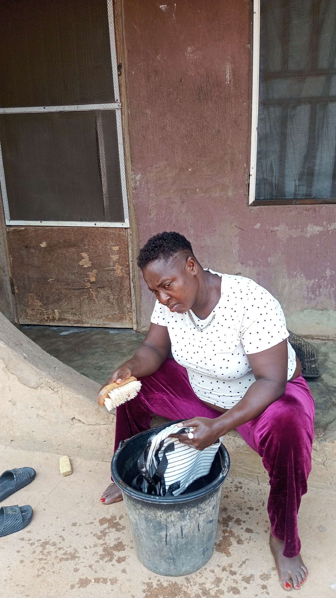In Onitsha, Nigeria, a woman sits outside a building, washing clothes in a plastic bucket. She holds a scrub brush in her right hand, and in the bucket, she is washing a striped garment. A bar of soap lies on the ground next to a pair of sandals. The building has a wooden door on the left and a window on the right. The woman wears a white polka dot top and dark purple pants. Her toes are painted red.
