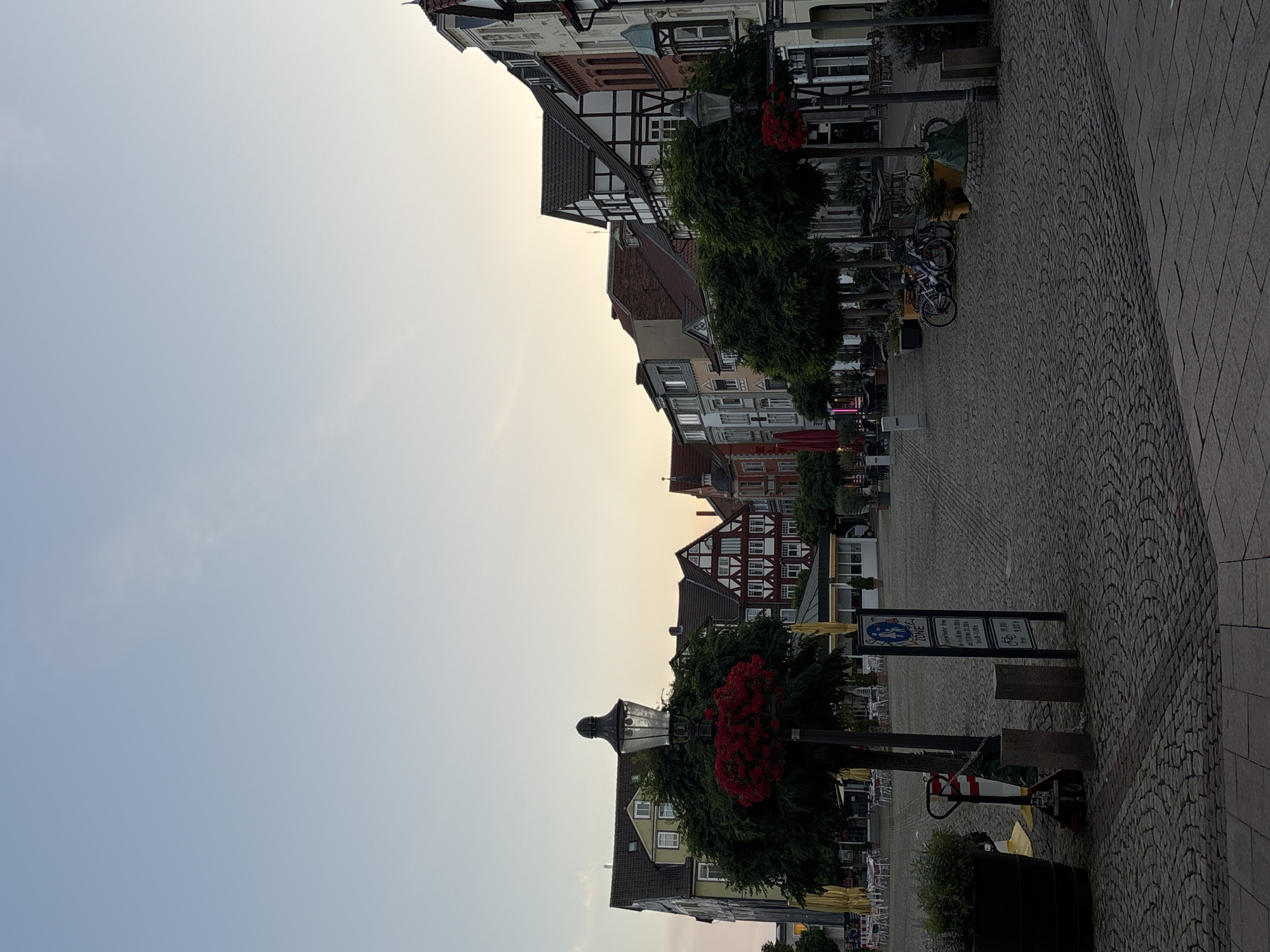 The image shows a cobblestone plaza in an unknown city. The plaza is lined with traditional European-style buildings, many featuring half-timbered architecture. In the foreground, there is a lamppost adorned with red flowers, a large potted plant, and a hand truck. Several trees are planted along the perimeter of the plaza, and outdoor seating areas with tables and umbrellas are visible in the background, suggesting a commercial or dining area. A few bicycles are parked near some of the trees. The sky is clear with a soft, pale hue, indicating either early morning or late evening. There are no people visible in the scene.