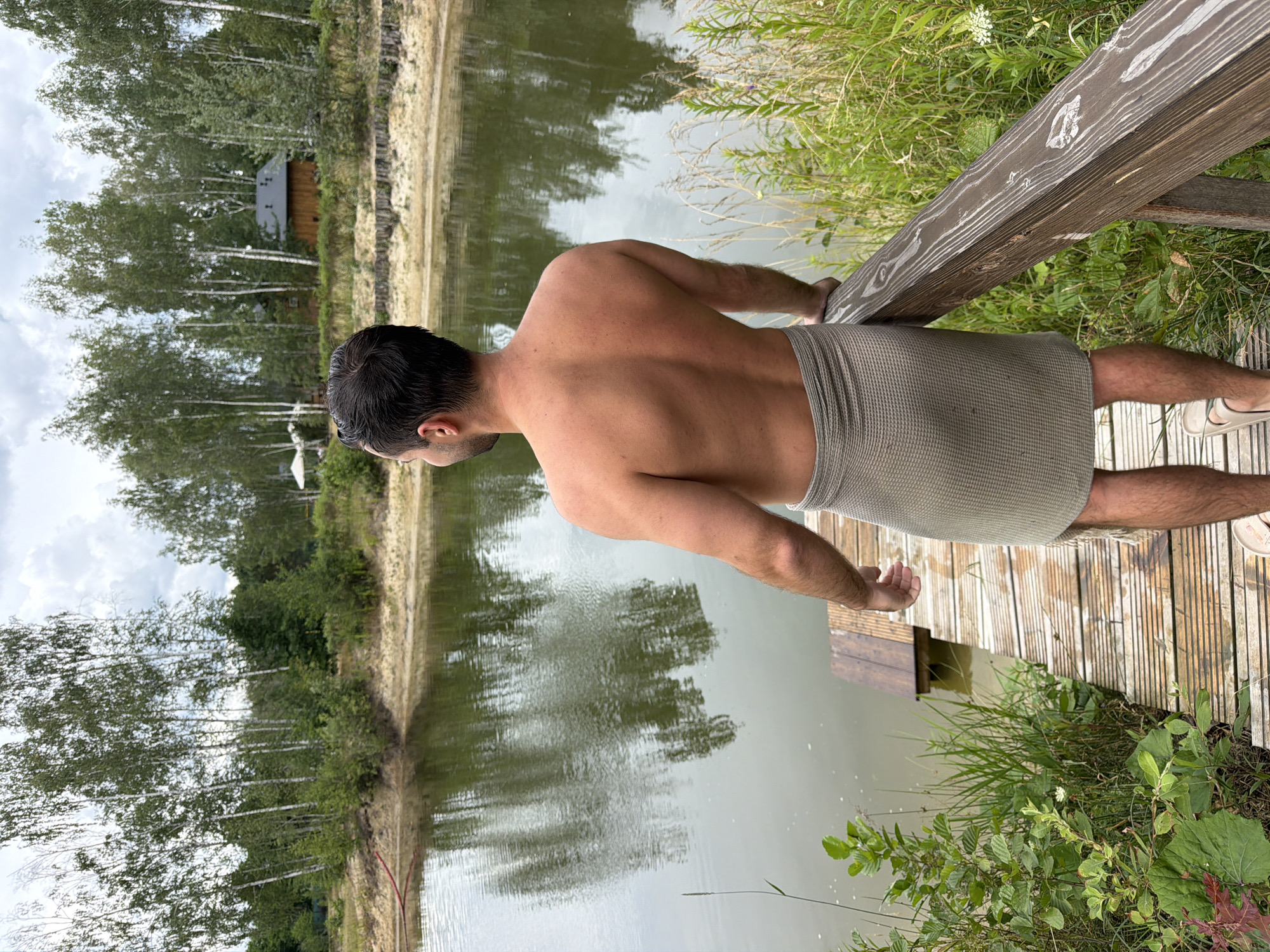 A man stands on a wooden dock, facing away from the camera, looking out at a calm lake. He is wearing a gray towel wrapped around his waist. The man has dark hair and is shirtless, revealing his back. The lake is surrounded by trees, with a small wooden building visible in the background. The sky is overcast. The scene appears to be a serene, natural setting. It is not possible to determine the exact location within Dobrzyń, Poland from the image alone.