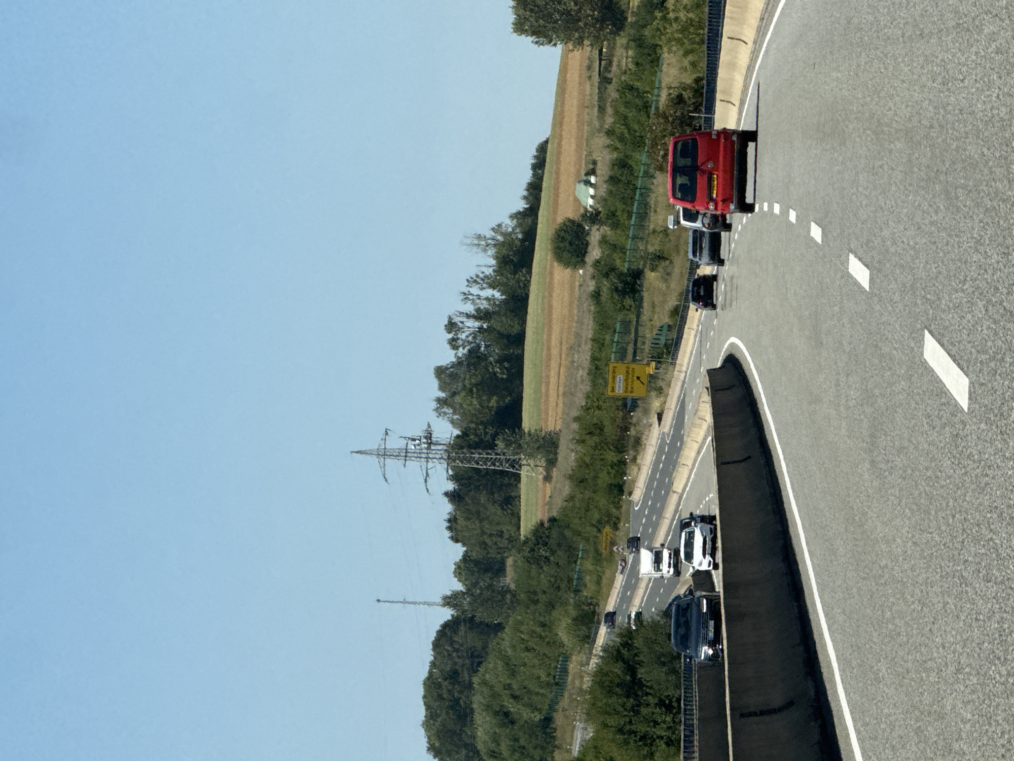The image captures a view from a highway in Germany, likely near Bad Lauterberg im Harz, given the yellow directional sign visible in the middle ground. The highway is multi-lane, with a concrete barrier separating the lanes. Several vehicles are present on the highway, including a red van in the foreground, a white truck, a dark SUV, and other cars. The surrounding landscape features rolling hills covered with green trees and fields, with a clear blue sky overhead. A tall electricity pylon is visible in the background, as is another, smaller tower. The overall scene depicts a typical sunny day on a German autobahn.