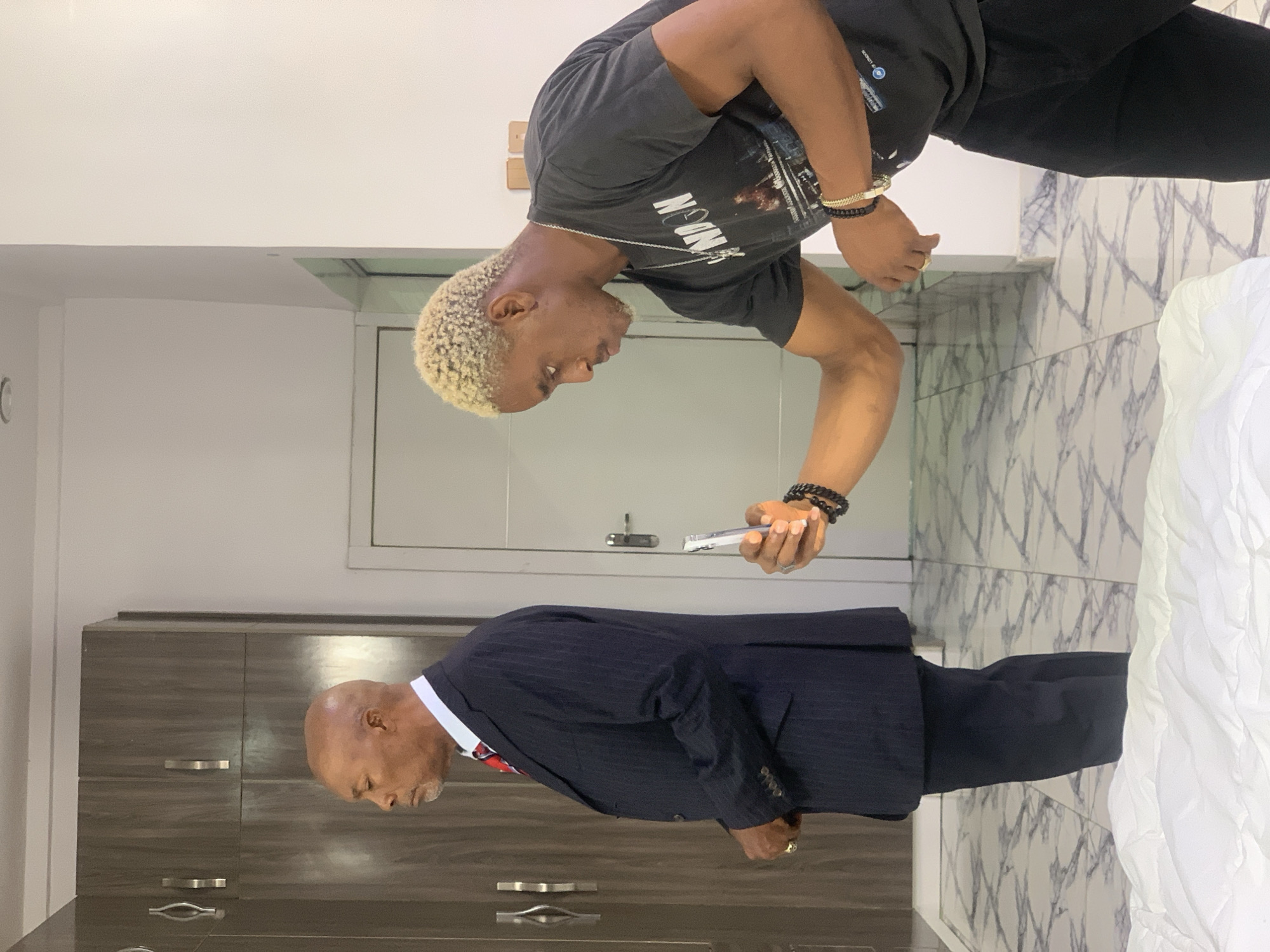 The image depicts an indoor scene in Lekki, Nigeria, featuring two Black males in what appears to be a modern residential environment.
On the left side of the frame, an older male stands in profile, facing left. He is bald with a short grey beard and is dressed in a dark navy blue pinstripe suit, a white collared shirt, and a red and blue patterned tie. A gold ring is visible on his left ring finger. His eyes appear closed or are directed downwards, and his hands are clasped in front of him, partially obscured by his suit jacket. Behind him are two dark brown wooden cabinets or wardrobe units with silver handles.
To the right, a younger male is in a crouched, forward-leaning posture, actively holding a silver smartphone in his left hand, aimed towards the older man. He has short, bleached blonde curly hair, a light beard, and is wearing a dark grey t-shirt with a graphic print, black trousers, a silver chain necklace, and multiple bracelets on both wrists (black beaded, gold-colored, and a chunky black chain-link). A gold-colored ring is visible on his right middle finger. His gaze is fixed on the smartphone screen, indicating he is operating the device, likely for photography or video recording.
The floor is covered with white and grey patterned tiles featuring an abstract, marbled design. The walls are predominantly white. A white door with a silver handle is visible in the background between the two individuals. A large, white quilted duvet or blanket occupies the lower foreground of the image. The scene is illuminated by consistent indoor lighting.