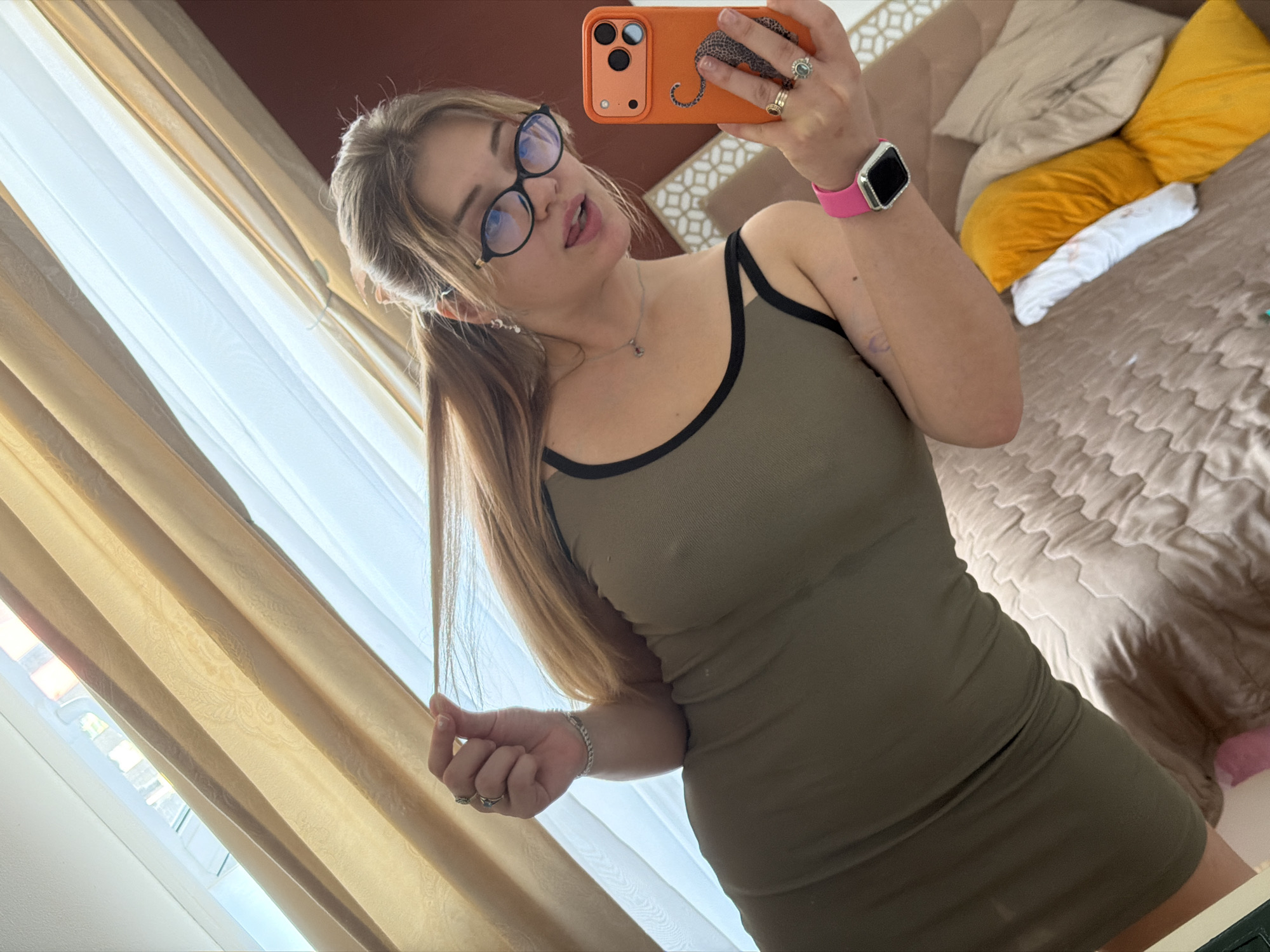 A young woman stands indoors, holding an orange phone in her right hand to take a selfie. She has long blonde hair styled in pigtails and is wearing black-rimmed glasses. She is wearing a form-fitting olive green dress and a pink smartwatch on her left wrist. The background includes light-colored curtains and a bed with beige bedding and yellow pillows. The location appears to be a residential interior in Большая Шильна, Russia.