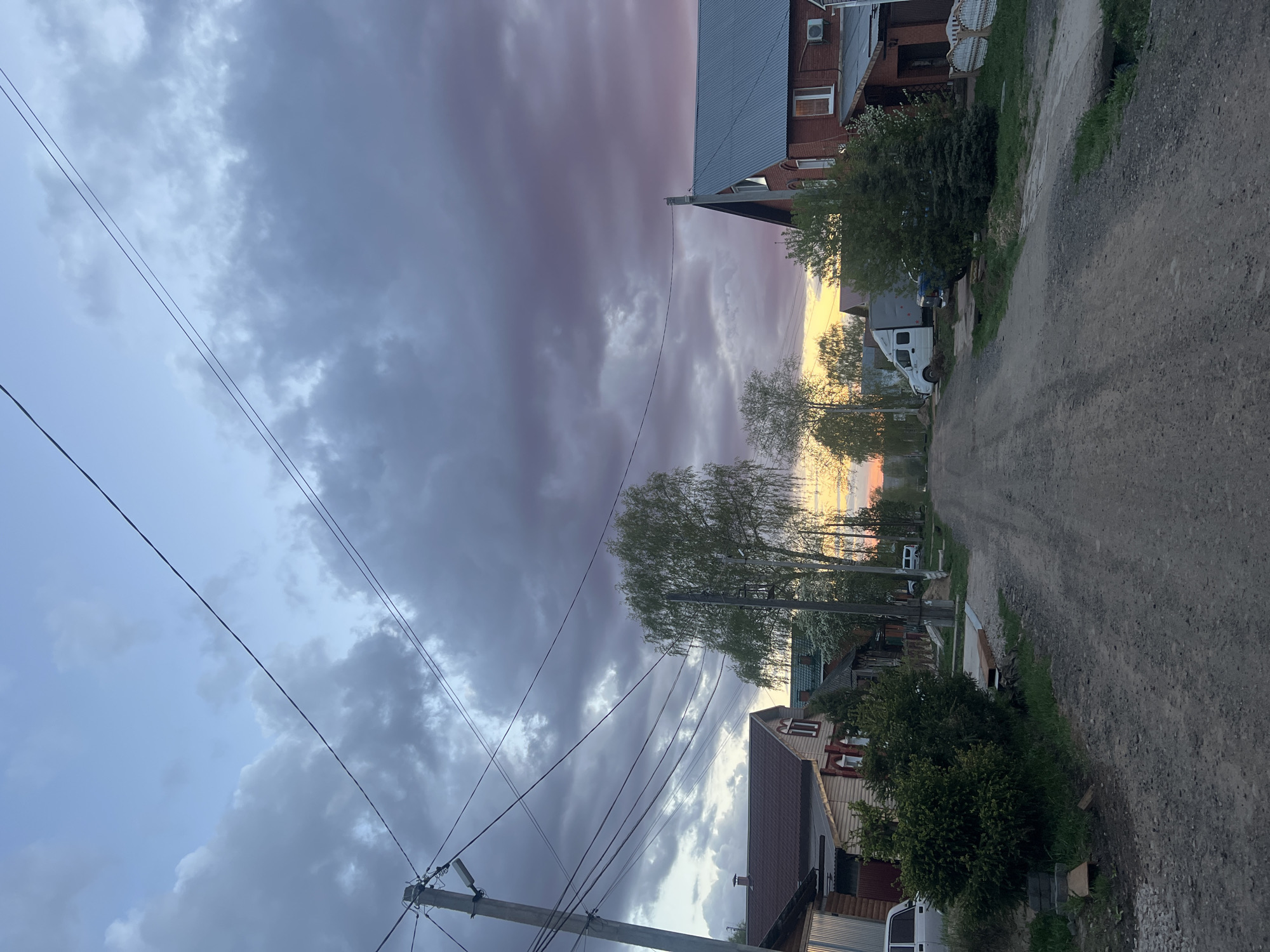 The image depicts a gravel road in a residential area, likely in Votkinsk, Russia based on the image context. The road stretches into the distance, flanked by houses and trees on both sides. Overhanging power lines crisscross the view. The sky is filled with dramatic clouds, displaying hues of grey, blue, pink, and yellow, suggestive of either sunset or sunrise. There are some vans parked along the street. No people are visible in the image.