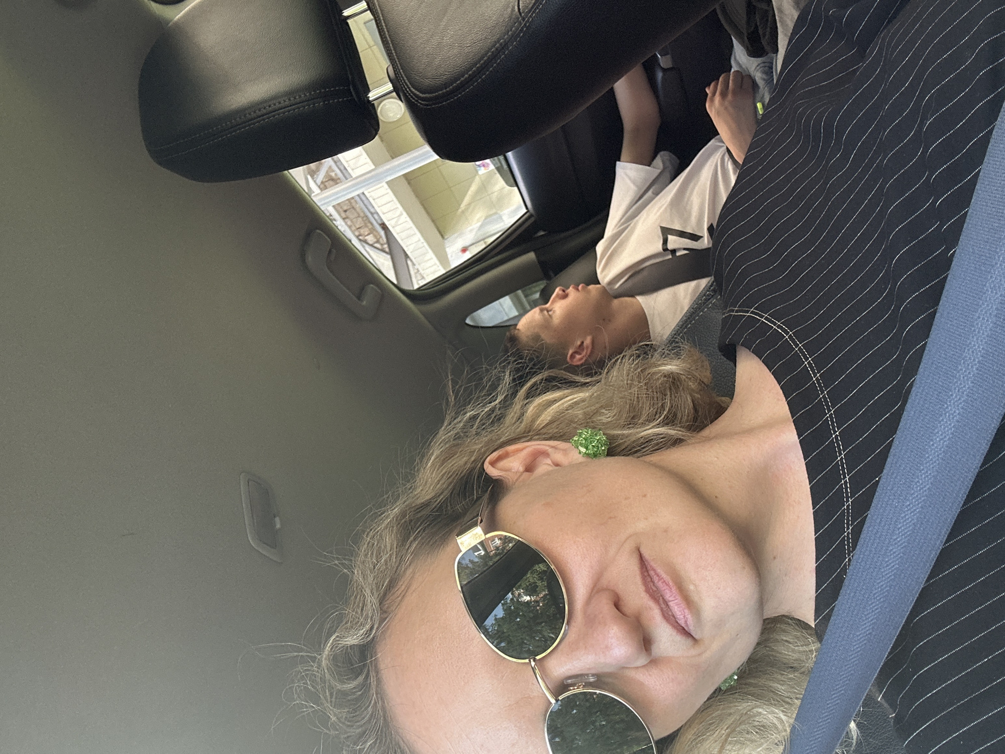 The image shows a selfie taken inside a car. A woman wearing sunglasses and a striped jacket is in the foreground, secured with a seatbelt. In the background, a child is visible, possibly lying down or reclined in a seat. The car's interior features black leather headrests and a light-colored ceiling. The reflection in the sunglasses shows a building exterior with a railing, indicating the car is near structures. The exact location within Izhevsk, Russia, is not discernible from the image.