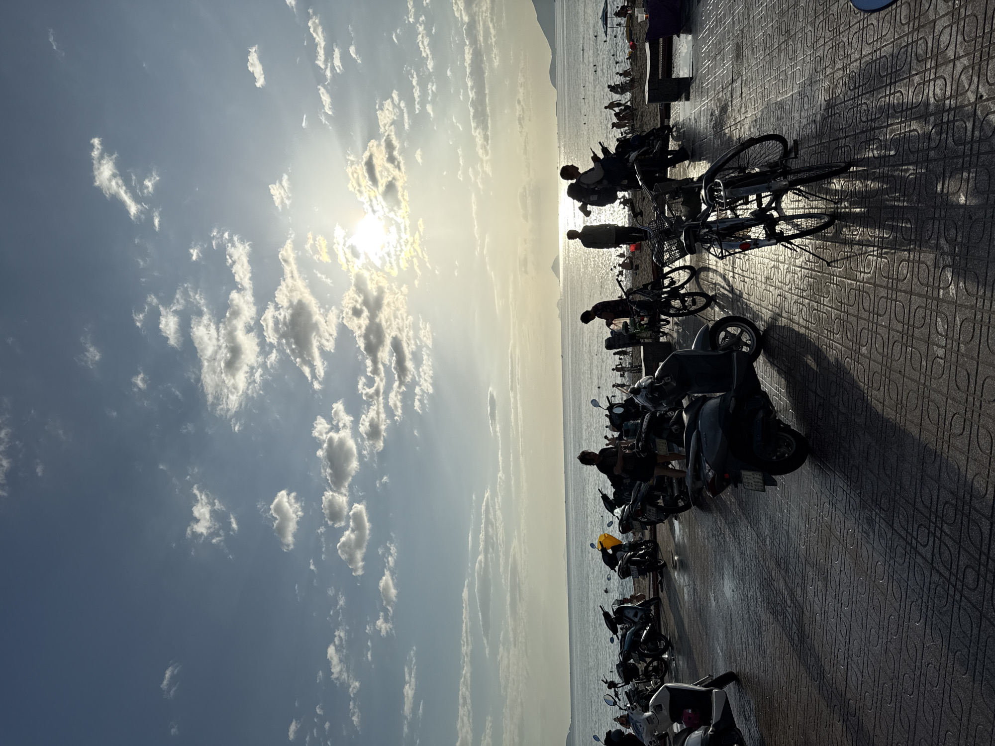 A silhouetted scene at a beach promenade in Nha Trang, Vietnam, features a paved walkway in the foreground with parked motorcycles and bicycles casting long shadows. The sun, partially obscured by clouds, is setting or rising over the ocean, creating a bright glow and illuminating the sky with scattered white clouds. Beyond the walkway, a beach is populated with numerous people, many of whom are also silhouetted. The ocean stretches to the horizon, where distant landmasses are visible. The overall atmosphere is serene and evocative of a popular coastal destination.