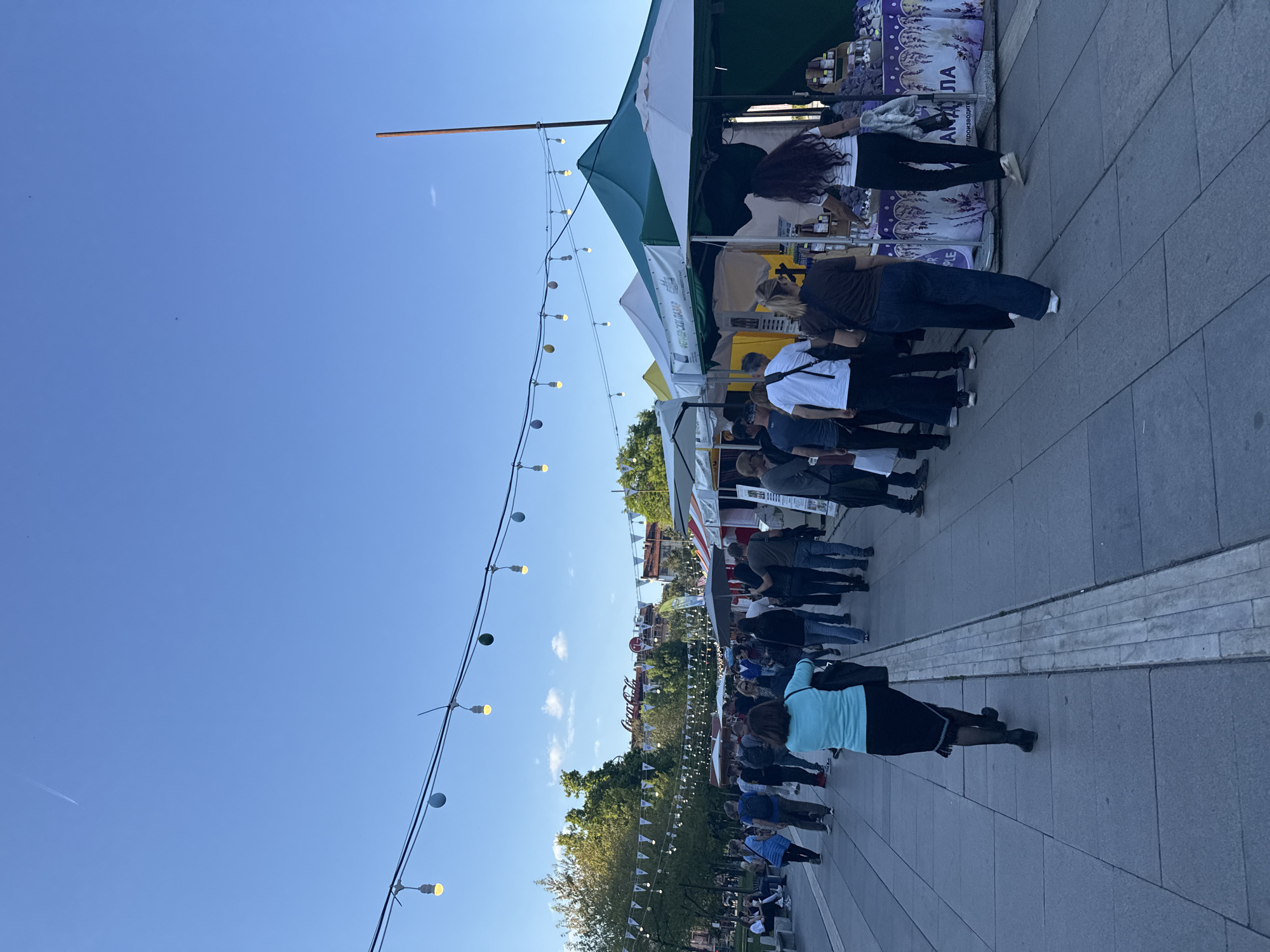 The image shows a bustling street scene, potentially a fair or market, in Sofia, Bulgaria. There are numerous people strolling along a paved area, with vendor tents visible on the right side of the frame. The tents are various colors, including green and white. Many people are milling around and looking at the vendor tents, while others are standing in line. String lights with decorative bulbs are strung above the crowd. In the background are trees and buildings, suggesting a city center location. The sky is a clear blue.
