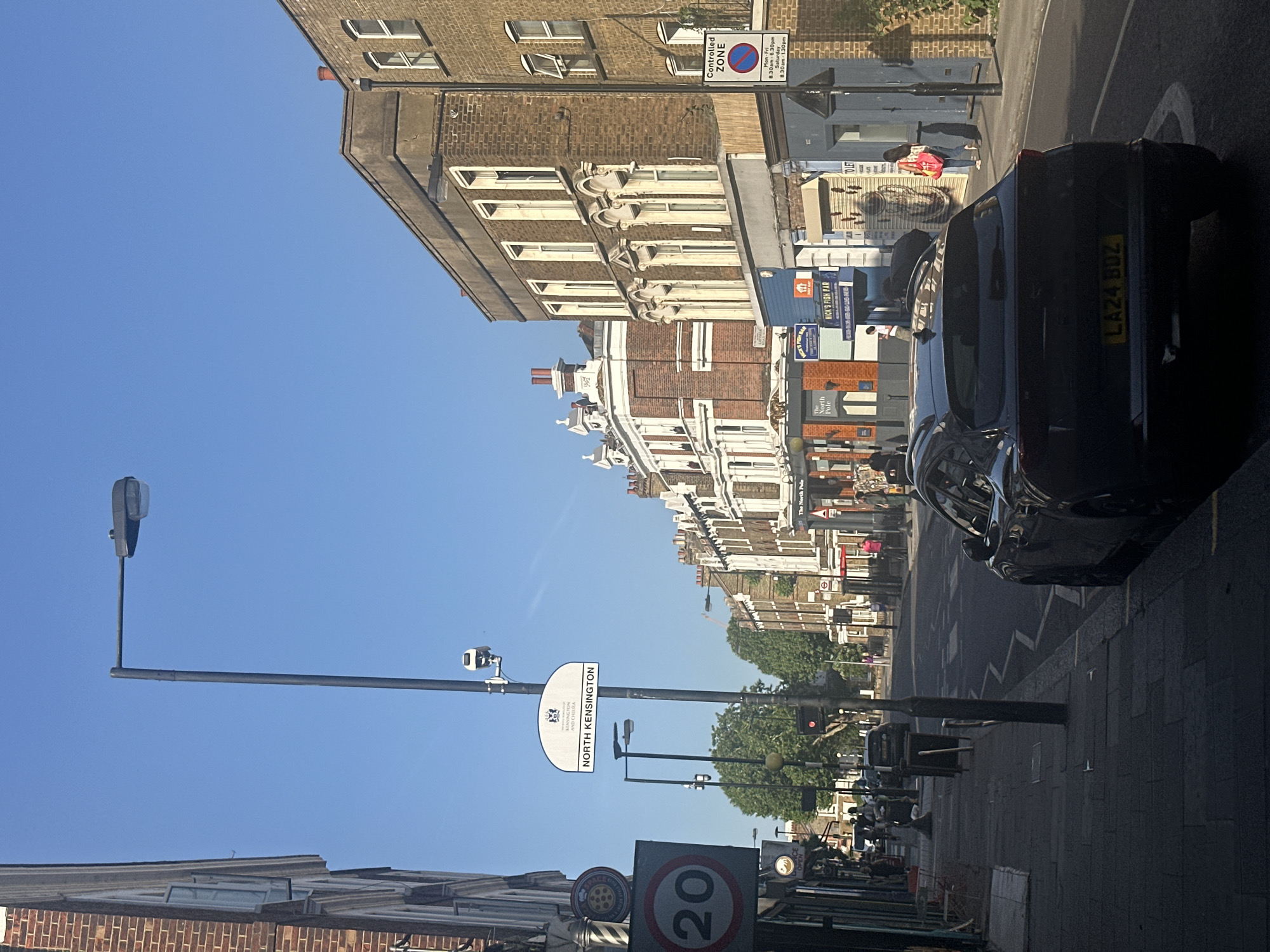 The image captures a street scene in North Kensington, London, United Kingdom. The view is oriented along a street lined with buildings of varying architectural styles. A black car is parked on the right, its license plate visible. Street signage indicates "North Kensington" and a "20" speed limit. A traffic light is seen further down the street, and a "Controlled Zone" parking sign is visible on a building. The buildings appear to be a mix of residential and commercial, with visible storefronts including "Mick's Fish Bar" and "The North Pole". People are seen walking near the shops. The sky is clear and blue.
