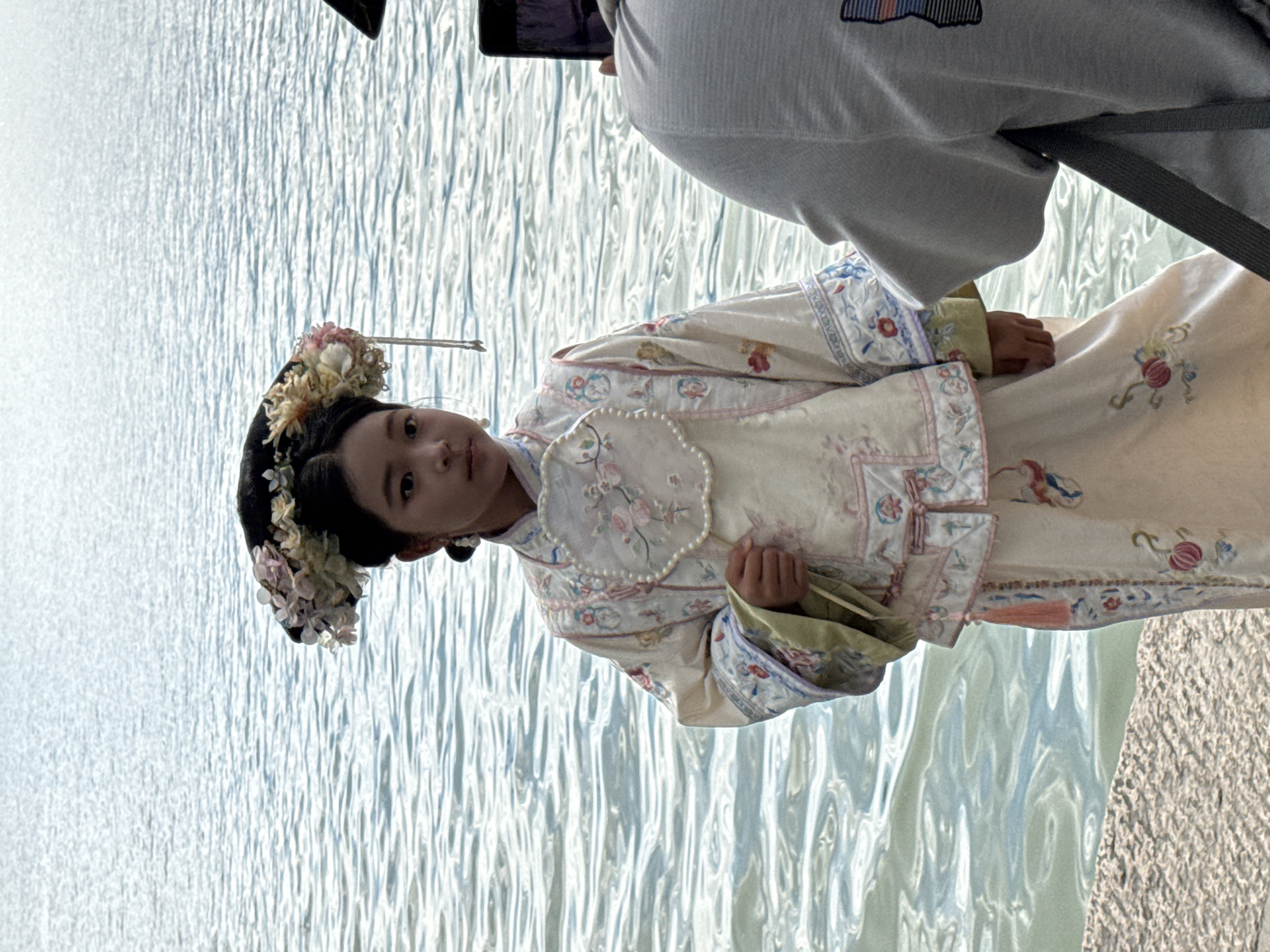 An outdoor scene in Haidian District, China, captures a young girl in traditional attire being photographed beside a body of water. The central figure is a young East Asian girl, standing upright and looking directly forward. She wears an elaborate traditional Chinese costume, featuring an off-white long-sleeved top and matching long skirt, both richly embroidered with floral patterns in various shades of pink, blue, green, and red. The top has a round collar and a distinctive fan-shaped detail on the chest, adorned with a string of small white pearls. Pearl drop earrings are visible on her ears. Her intricate black hairstyle is topped with a large, ornate headdress composed of numerous light-colored artificial flowers (white, pink, yellow, purple), with a delicate string of beads or pearls dangling from one side. In her right hand, she holds a partially visible, folded fan. To the right of the frame, the back and right shoulder of an adult individual are visible. This person wears a light gray short-sleeved top with a partial graphic design on the back and holds a dark-colored smartphone horizontally, pointing it towards the girl, presumably taking a picture or video. A dark strap, possibly from a bag, crosses their back. The background consists of a vast body of water, likely a lake or a wide river, with a rippled surface that reflects the bright daylight, appearing light blue-grey. The subjects are standing on a textured, light-colored stone or concrete surface, possibly a pathway or pier, visible in the foreground. The scene is illuminated by natural daylight.