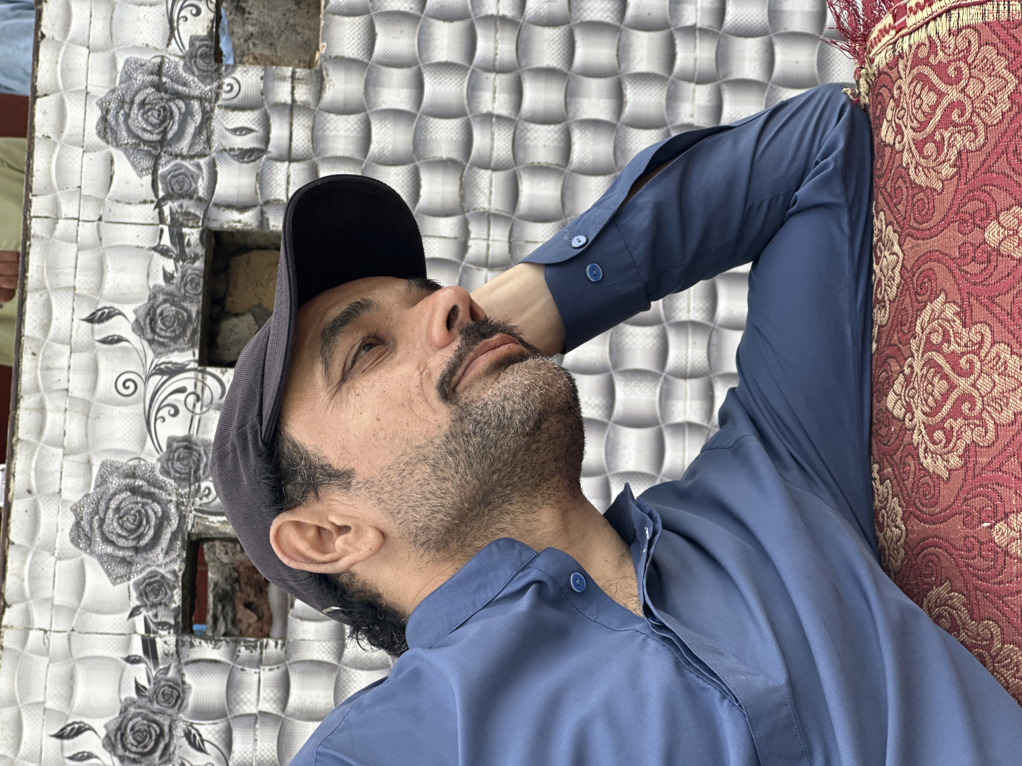 A man is depicted reclining or resting, with his head tilted back and his eyes closed or looking upwards. He appears to be in a state of relaxation. He has a dark beard and mustache and is wearing a dark blue long-sleeved collared shirt, buttoned up, and a black baseball cap. His right arm is bent, resting above his head, while his body is partially obscured by a decorative red cushion with intricate gold embroidery on which he is leaning.
The background features a tiled wall with a dual pattern. On the left, some tiles are adorned with elegant gray rose designs and swirling patterns. Adjacent to these, and extending across the background, are tiles with a textured, possibly geometric or wavy relief pattern in shades of gray and white. There is a visible gap or broken section in the wall near the man's head, revealing what appears to be rough construction materials or bricks within. The lighting is bright, suggesting daytime. No discernible text is visible in the image.