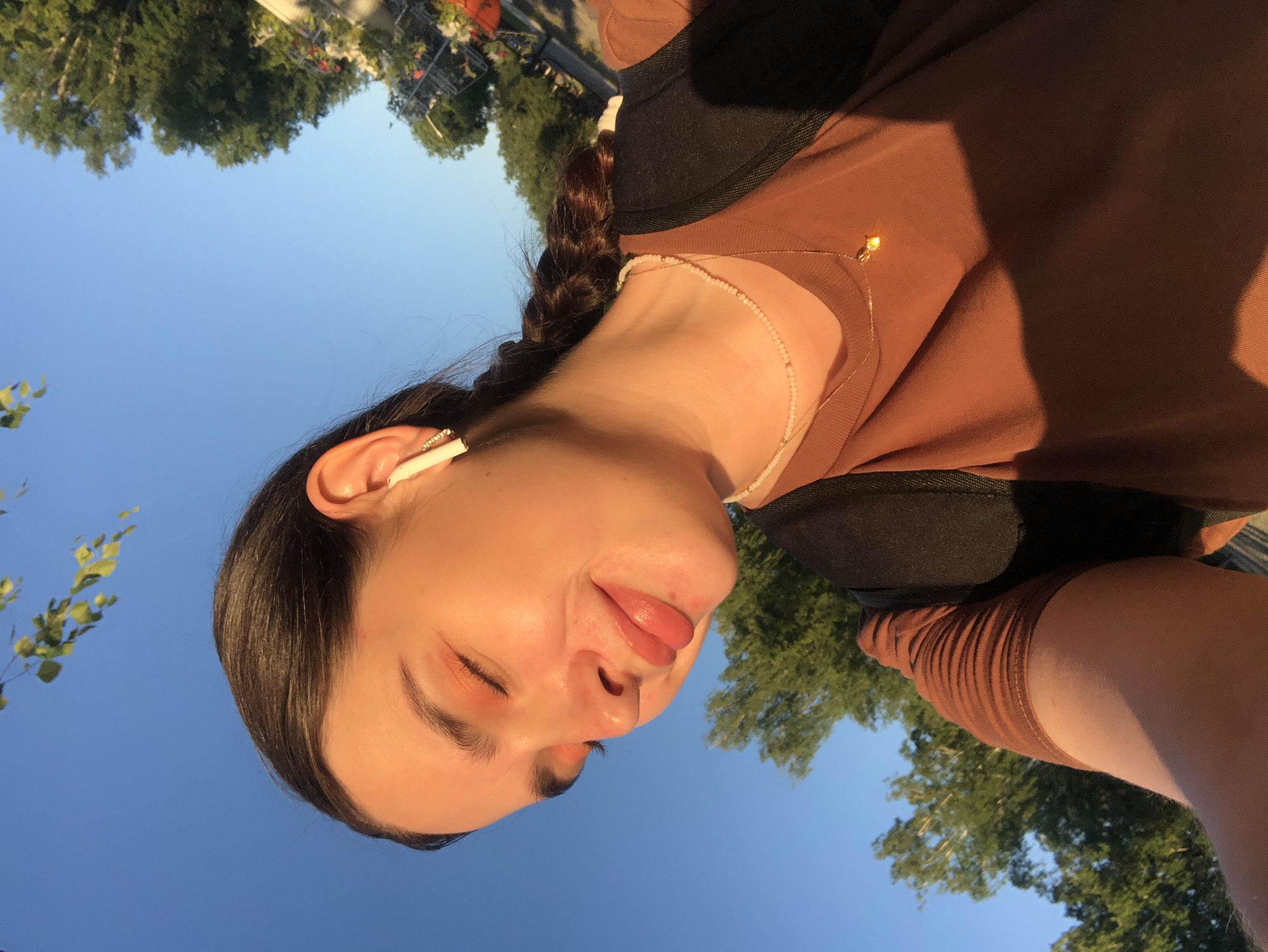 The image is a close-up, low-angle selfie of a young woman with her eyes closed and a slight smile. She has dark hair pulled back into a braid and is wearing a brown t-shirt, a black backpack, and a delicate gold necklace with a pendant. She also has a white earbud in her right ear. The lighting suggests it is late afternoon or early evening, with warm sunlight illuminating her face. The background is a clear blue sky with some green trees visible on the right side, suggesting an outdoor setting in Chaikovsky, Russia.