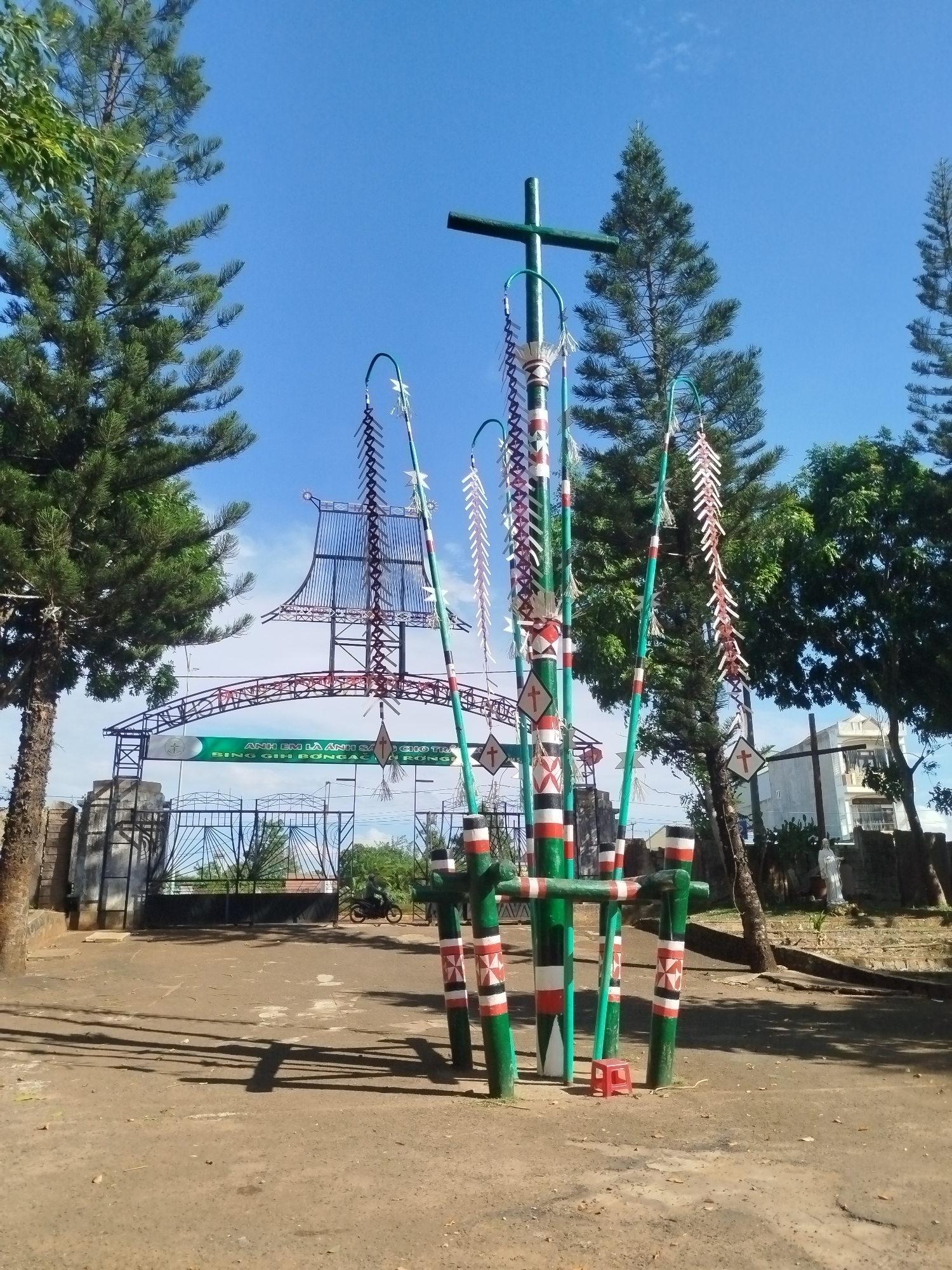 This image depicts the entrance to a site in An Phu Ward, Vietnam. In the foreground stands a decorative structure comprised of painted poles and a cross at the top, adorned with hanging decorations. Behind it is an ornate gate with a roof resembling a traditional Vietnamese boat or house, featuring an inscription. In the background, a motorcycle is visible on the road within the site, and there is a hint of buildings and foliage beyond the gate. Tall pine trees and other greenery frame the scene, set against a clear blue sky. The ground is dirt or gravel.