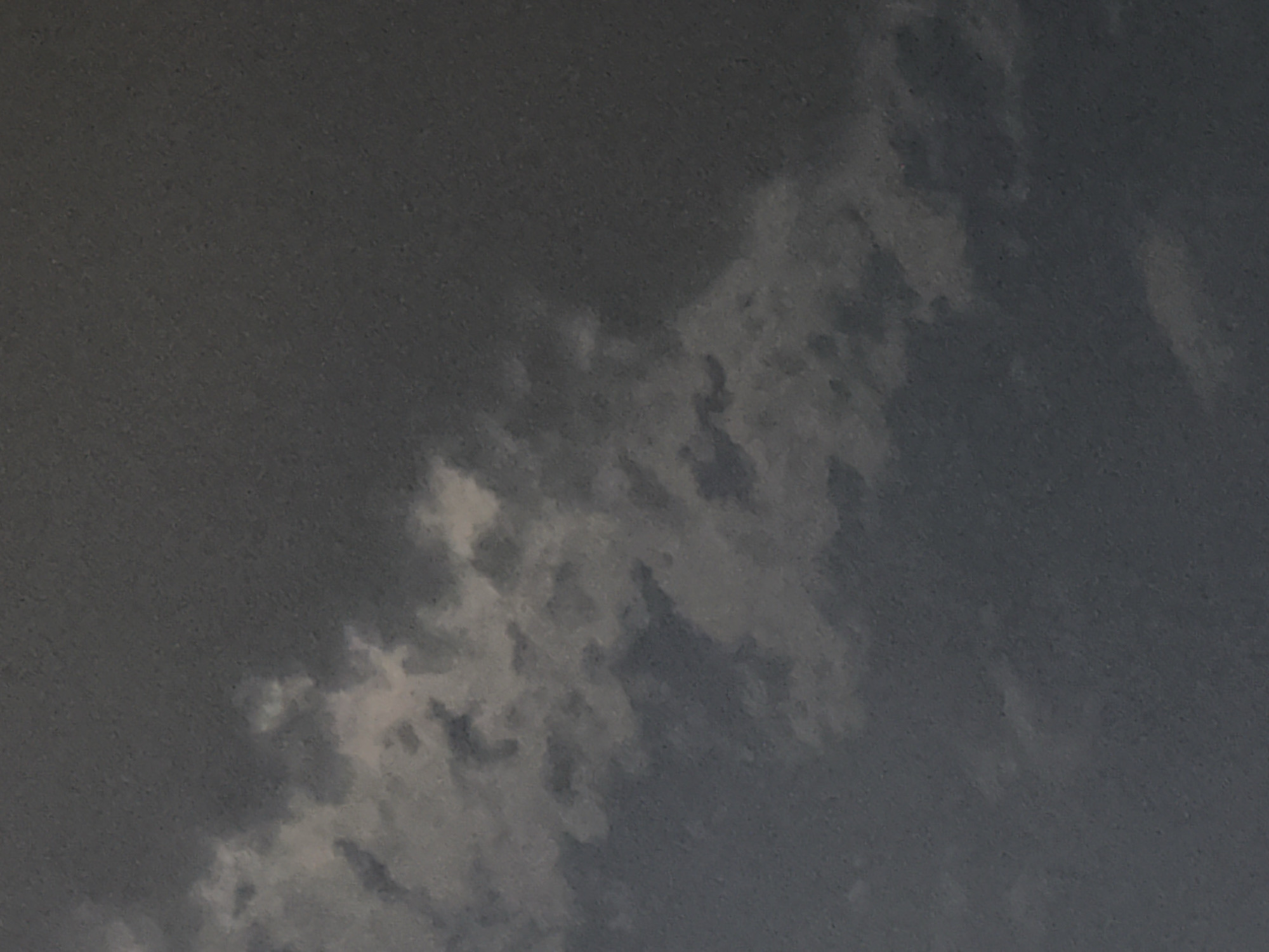 The image shows a close-up view of a cloudy sky. The clouds are predominantly dark gray, with some lighter, illuminated patches suggesting the presence of sunlight breaking through. The texture of the sky appears grainy, possibly due to image quality or atmospheric conditions. There are no discernible landmarks or elements that would indicate a specific location within the city of Minsk, Belarus. The image solely captures a segment of the sky.