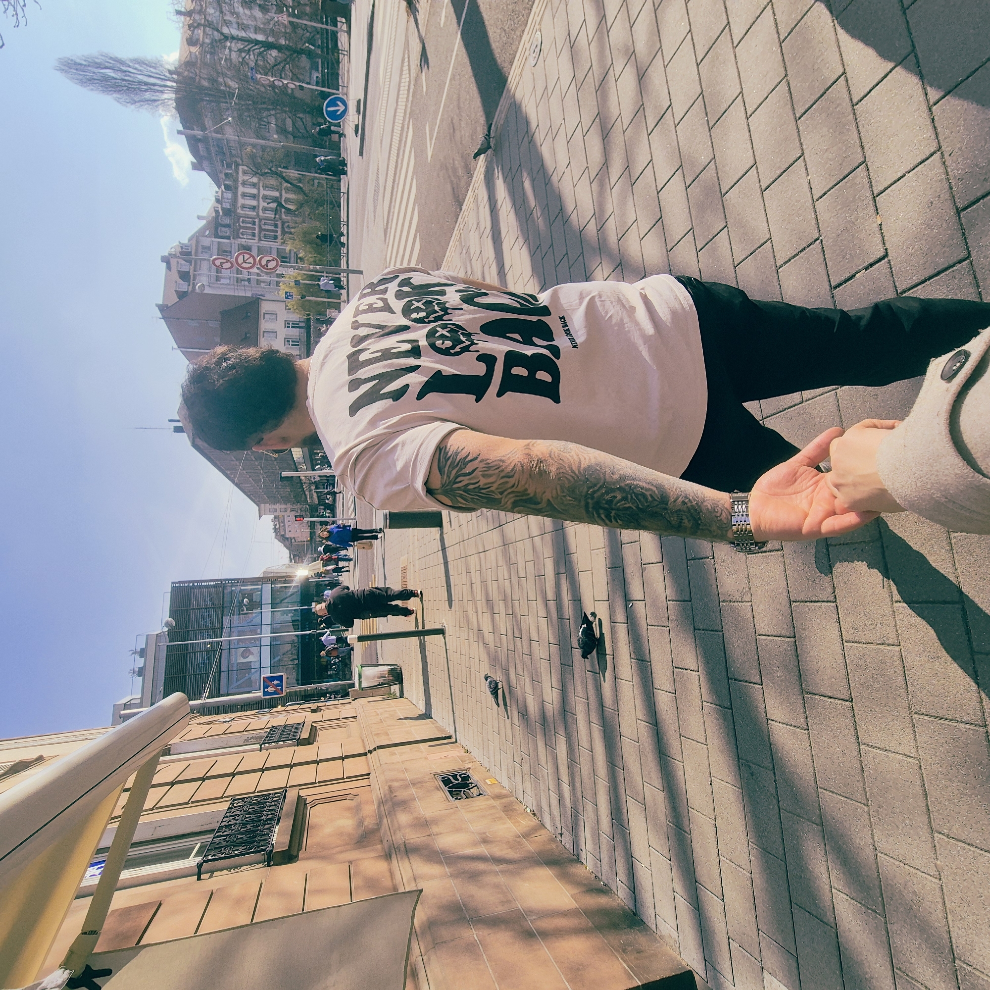 The image captures a first-person perspective on a sunny day in Strasbourg, France, specifically near Rue du Maire Kuss, with the Centre Commercial Place des Halles visible in the background. In the immediate foreground, a person's left hand, clad in a light beige coat, holds the right hand of a male individual. This individual, seen from behind, is walking forward on a brick-paved sidewalk. He has dark hair, wears a light-colored t-shirt with the black text "NEVER LOOK BACK" and two melting smiley face graphics, dark pants, and a full-sleeve tattoo on his left arm, accessorized with a two-tone metallic watch on his left wrist.
To the left, a multi-story building with a light brown stucco facade features windows with ornate black iron railings and a partially extended light-colored awning, casting shadows on the pavement. Several pigeons are present on the sidewalk. To the right, a wide street with a pedestrian crosswalk and various traffic signs, including a blue circular sign with a downward white arrow and red circular "no entry" signs, leads into the background. Multiple pedestrians are visible walking in the middle ground and background, dressed in casual attire. Architectural elements in the background include a modern glass and steel structure (Centre Commercial Place des Halles) on the left, and older European-style buildings with sloped roofs and dormer windows on the right, under a clear blue sky with minimal cloud cover.