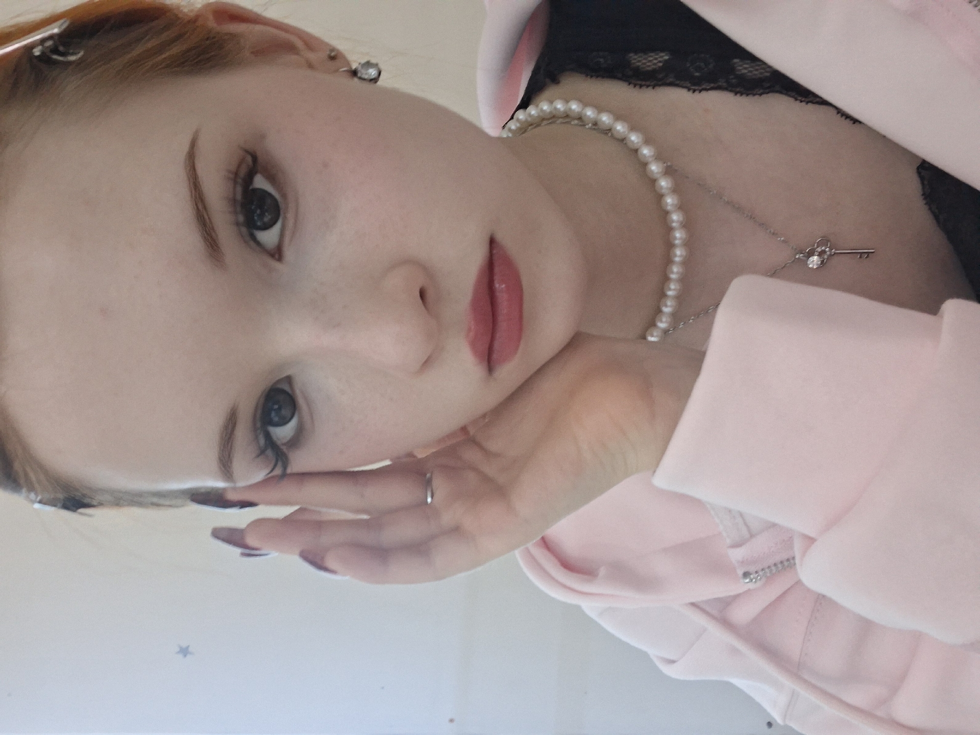A young woman with fair skin and red hair is posed in a close-up, slightly angled shot. She is wearing a pearl necklace with a silver key pendant, a black lace top, and a light pink jacket. Her hand is resting on her face. She has a ring on her finger. The reflection in the background suggests a flat surface, possibly a table or floor. The location is not evident from the image, so I cannot provide information about Chaikovsky, Russia.