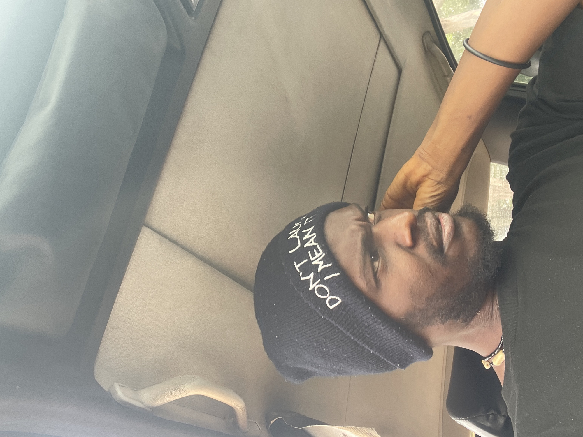 A man with dark skin and a beard is sitting in what appears to be a car. He's wearing a black beanie with white lettering that reads "DON'T LAUGH, I MEAN IT". He is also wearing a black shirt and a necklace. A black bracelet is on his arm. The interior of the car is visible, with beige upholstery. The scene is lit with natural sunlight. There is no information in the image to confirm the location is in Jos, Nigeria.