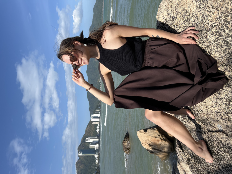 A female individual is seated on sun-drenched, textured light-colored rocks along a coastline. She is positioned towards the right of the frame, with her body angled leftward and her head turned upwards and to the right, exhibiting a smile. Her dark hair is pulled back, with loose strands around her face, and she wears brown-tinted sunglasses, which she holds with her right hand near her temple. She is dressed in a black ribbed halter-neck top and a loose, long, dark brown skirt with an elastic waistband. A dark beaded bracelet is visible on her right wrist, and a light-colored, teardrop-shaped earring is on her left ear. Her feet are bare.
In the midground, the sea, with a greenish hue, extends behind her, displaying small waves breaking near the shore and around several partially submerged dark rocks. The background features a clear blue sky with scattered white cumulus clouds. Further in the distance, across the bay, a city skyline composed of multiple high-rise buildings is visible at the base of lush green mountains. This setting, characterized by the specific coastal city architecture, mountainous backdrop, and bay, identifies the location as Nha Trang, Vietnam.