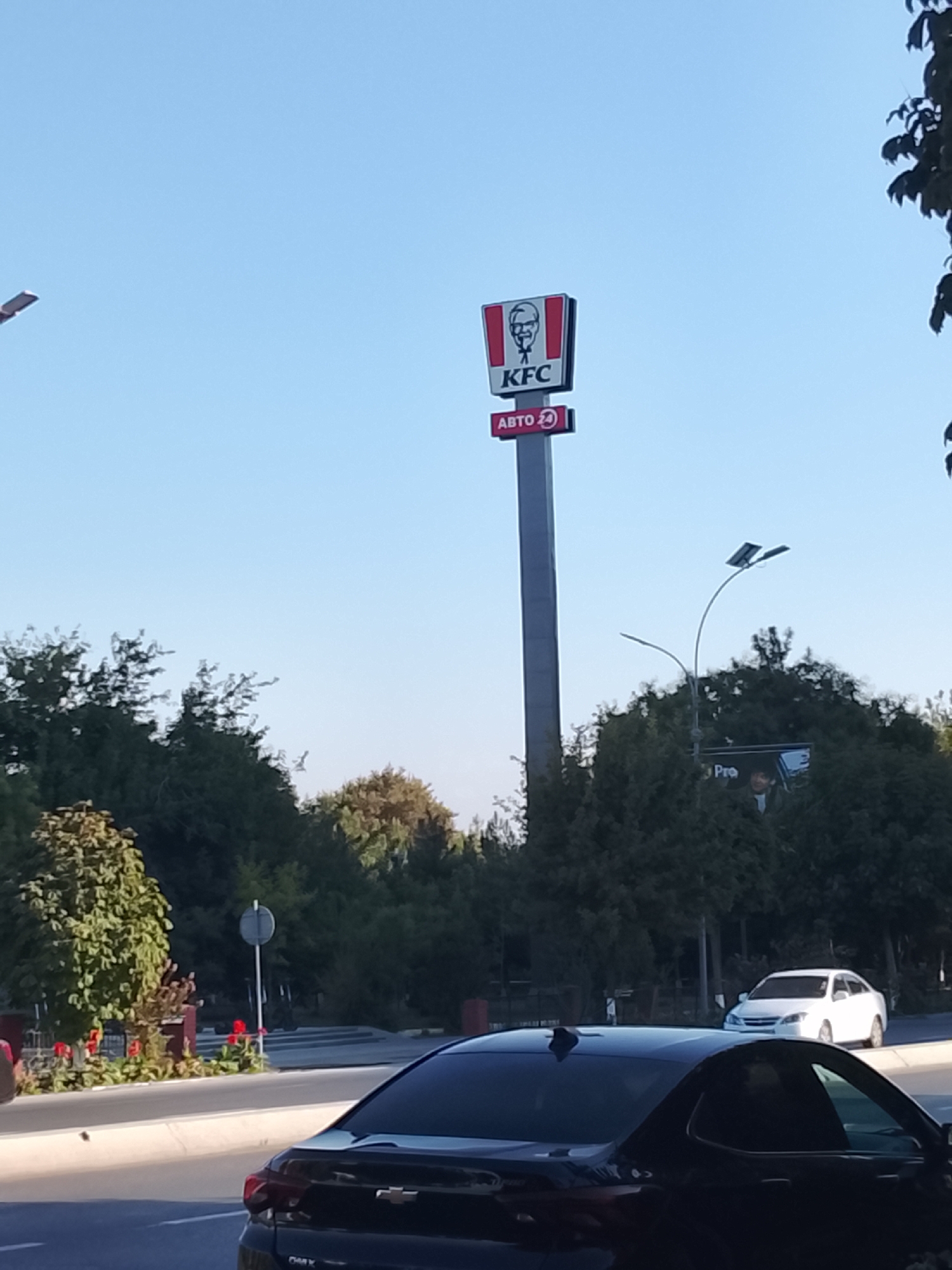 In Fergana City, Uzbekistan, a tall KFC sign stands prominently against a clear blue sky. The sign features the iconic KFC logo with Colonel Sanders' face and the text "KFC" in white letters on a red background, positioned above a red rectangular sign with "ABTO 24" written in white. Below the KFC sign, a street lamp with two solar panels is visible, curving elegantly over a backdrop of lush green trees. In the foreground, the rear of a dark-colored Chevrolet sedan is parked on a paved road, and a white sedan is parked further down the street. The scene is set during daylight hours, with the sun casting a bright illumination.