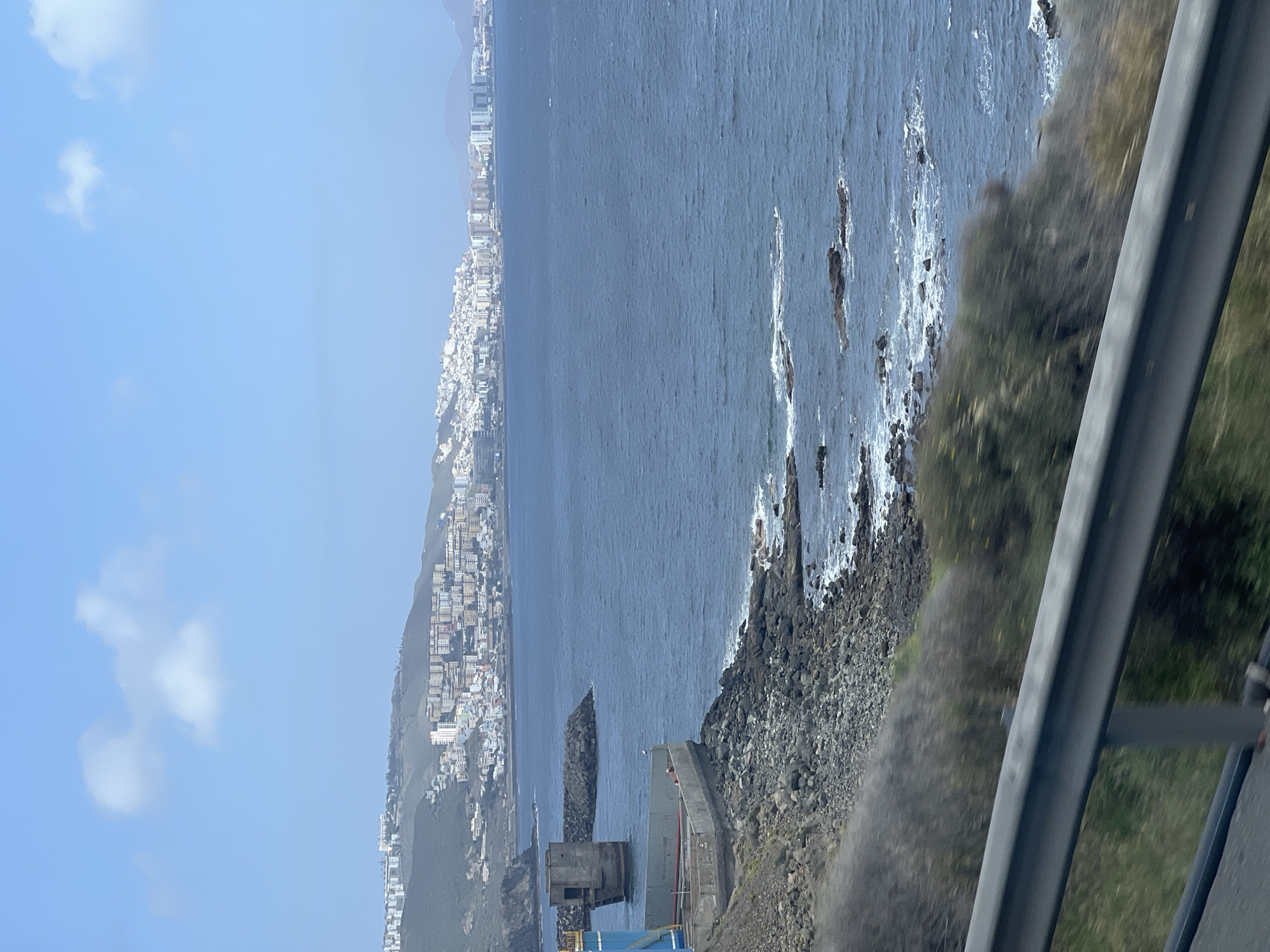 The image presents a panoramic coastal scene of Las Palmas, Spain, captured during a clear, sunny day. The dominant features include a vast ocean, a rocky shoreline, and a distant city sprawling along the coast.
In the upper portion of the frame, a bright blue sky stretches across, dotted with a few wispy white clouds. Below the sky, the deep blue waters of the Atlantic Ocean fill the midground, showing subtle textures from the movement of the water. Along the right side of the ocean, a rugged, rocky shoreline is visible, where white-crested waves crash against dark rocks, creating foamy spray. The land directly adjacent to the sea appears steep and covered with rough, dark rocks and patches of green and brown vegetation, suggesting a wild, natural coastline.
Further to the left, along the distant horizon, a dense urban landscape of Las Palmas extends horizontally. Numerous buildings, predominantly white and light-colored, are visible, climbing up the hilly terrain, indicating a large, developed city. Closer to the foreground on the left, a man-made structure, possibly a fortified breakwater or an old industrial building, is constructed from concrete and rock, jutting out into the water.
In the immediate foreground on the right, a dark gray guardrail, consistent with those found along roads or elevated viewing points, angles across the image. The foreground also shows blurred green and brown vegetation, suggesting the photograph was taken from a moving vehicle or a vantage point close to a road. The scene is devoid of any visible people or discernible text. The bright sunlight and clear sky indicate a pleasant daytime setting.