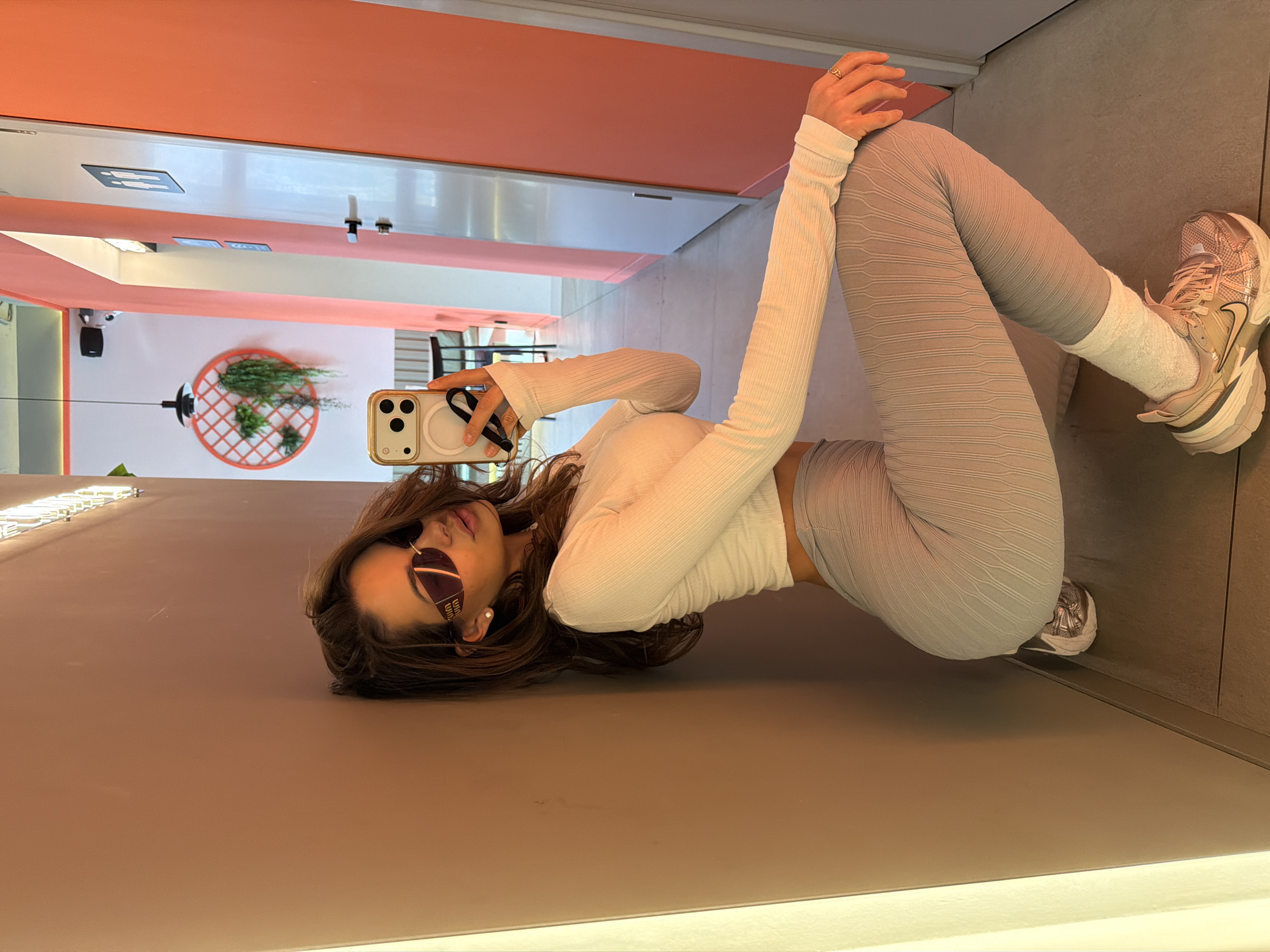 A woman wearing a white long-sleeved shirt, gray leggings, and sneakers is lying on the floor, taking a selfie with her phone. She is wearing sunglasses. The location is an indoor space with peach-colored walls and a white ceiling. In the background, there's a mirror reflecting a seating area and a decorative piece with plants. The wall and floor have a solid, neutral tone. The image does not provide sufficient information to determine if the location is in Krasnoyarsk, Russia.