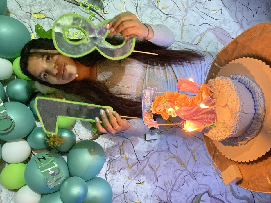 A young woman poses for her 18th birthday. She holds up number cutouts made of green glitter. The balloons, also green, and a tiered cake with a Rapunzel topper with lights are also in frame. The "Feliz Cumpleaños" banner on the cake indicates a birthday celebration. The background displays a patterned wallpaper.