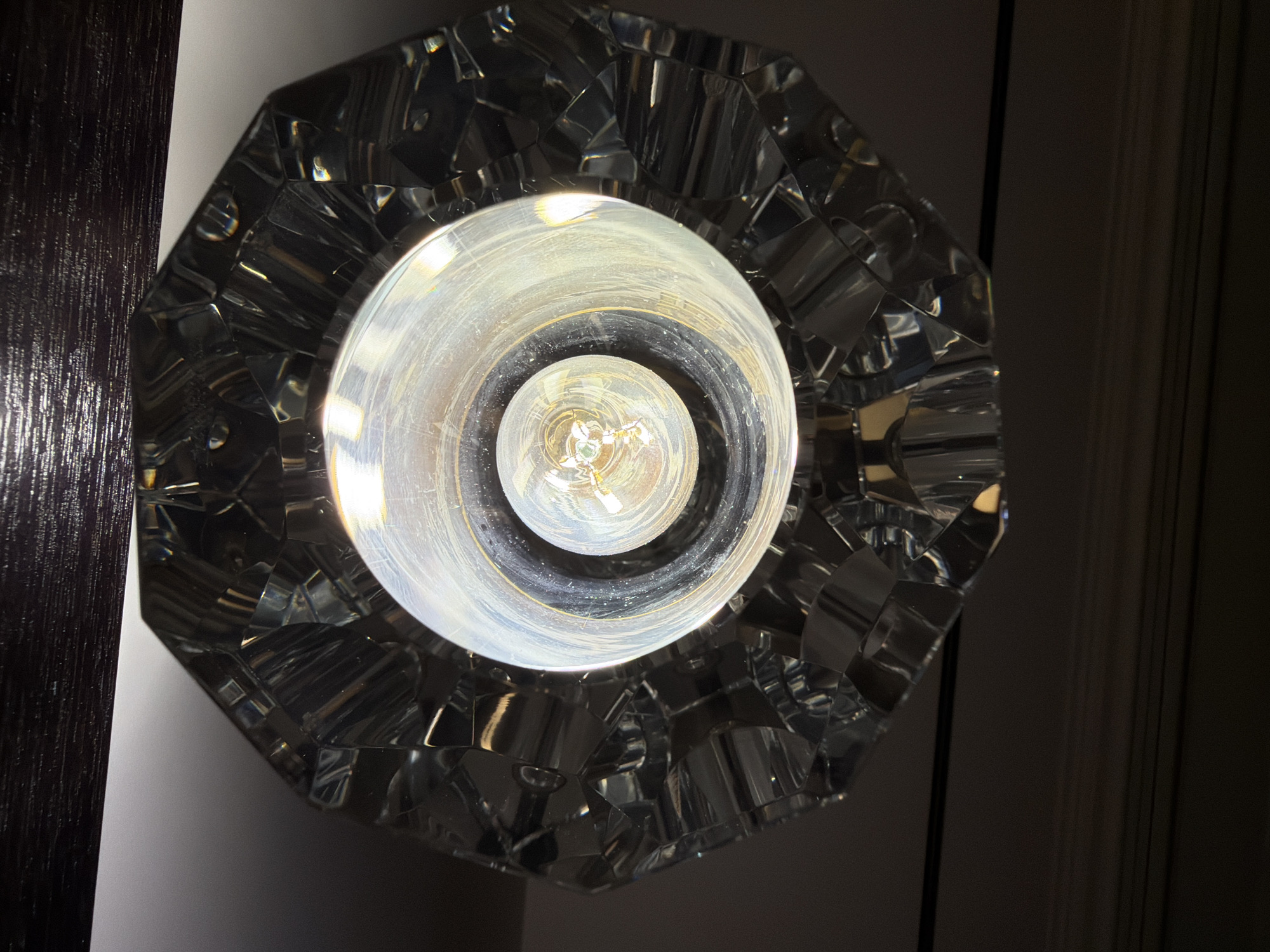 An interior, downward-facing, close-up shot shows a light fixture. The fixture has a faceted, clear glass exterior and a central glass globe surrounding a visible incandescent light bulb. The light bulb is illuminated, casting a bright, warm glow. The fixture is attached to a dark, textured surface, possibly a ceiling or beam. The background is dark and out of focus. The location of the image is not specified and cannot be determined.