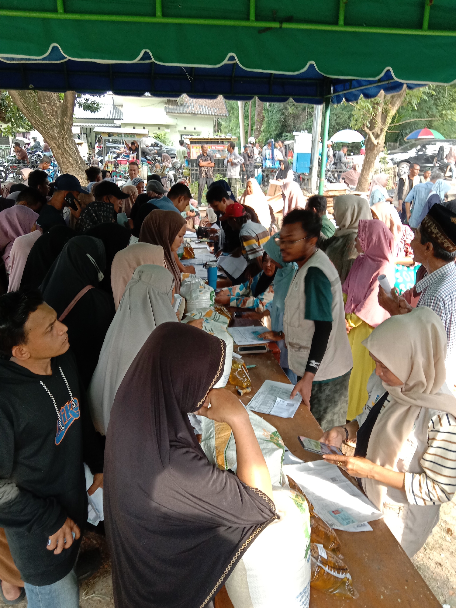 The image depicts a gathering of people under a green and blue awning, possibly in Lhokseumawe, Indonesia. Many individuals are wearing hijabs and other traditional clothing. They are congregated around a long wooden table where documents and packages are placed. Some people are handing over documents and receiving items from the table, and one woman is using a smartphone. In the background, there is a parking area with motorcycles and people milling about. Trees and buildings are also visible in the background. The scene appears to be an organized distribution event.