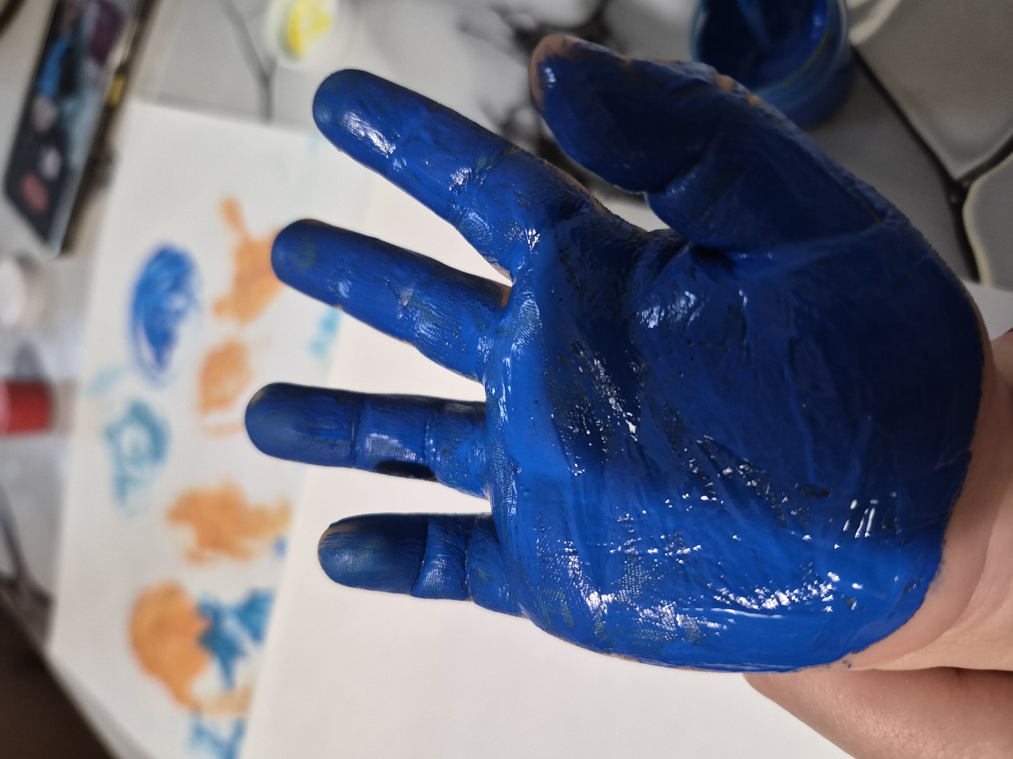 A hand, covered in blue paint, is held up in the foreground. The fingers are spread slightly apart. In the background, there is a white piece of paper with blue and orange paint smudges. There are also containers of paint, including red and yellow, and a smartphone visible in the upper left. A jar of blue paint is also visible in the background. The environment appears to be a domestic setting, possibly a home or art studio. There is no verifiable information to confirm that the photo was taken in Tobolsk, Russia.
