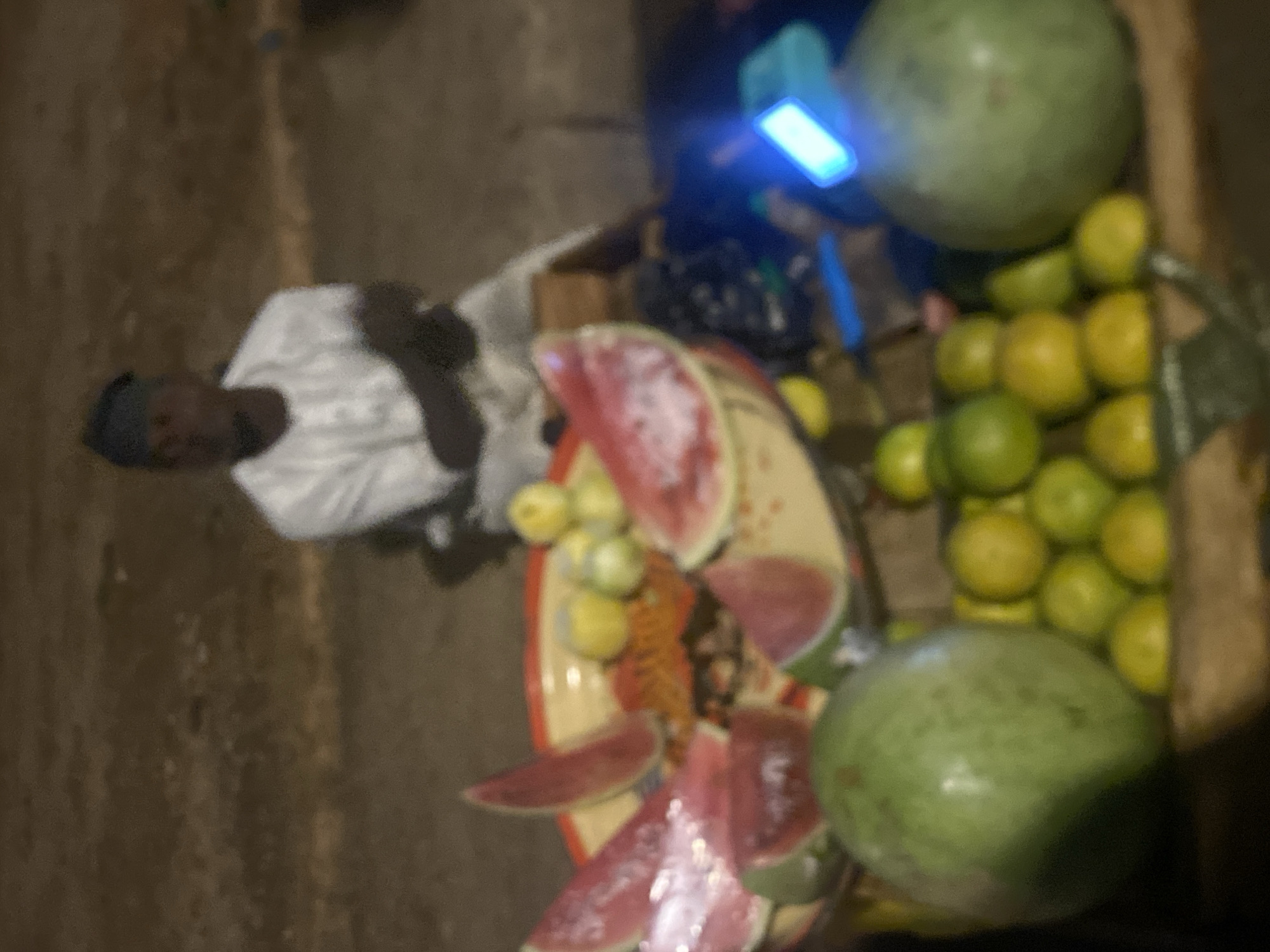 The image appears to depict a roadside fruit stand in Bukuru, Nigeria. A man, possibly the vendor, stands to the left. The fruit stand displays watermelons (whole and sliced), and a pile of green and yellow citrus fruits. A small blue light illuminates the stand. The surrounding environment is indistinct and appears to be outdoors, possibly a street.