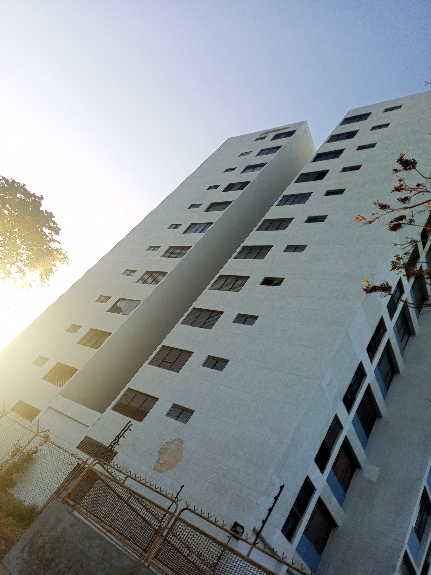 The image shows a low-angle shot of a tall, light-colored building divided into two sections with a narrow vertical gap in between. The building appears to be several stories high, featuring regularly spaced windows of varying sizes. A tree is visible on the left side, partially obscured by sunlight. A fence and gate are in the foreground. The sky is clear and bright. The location is Unknown, Unknown.