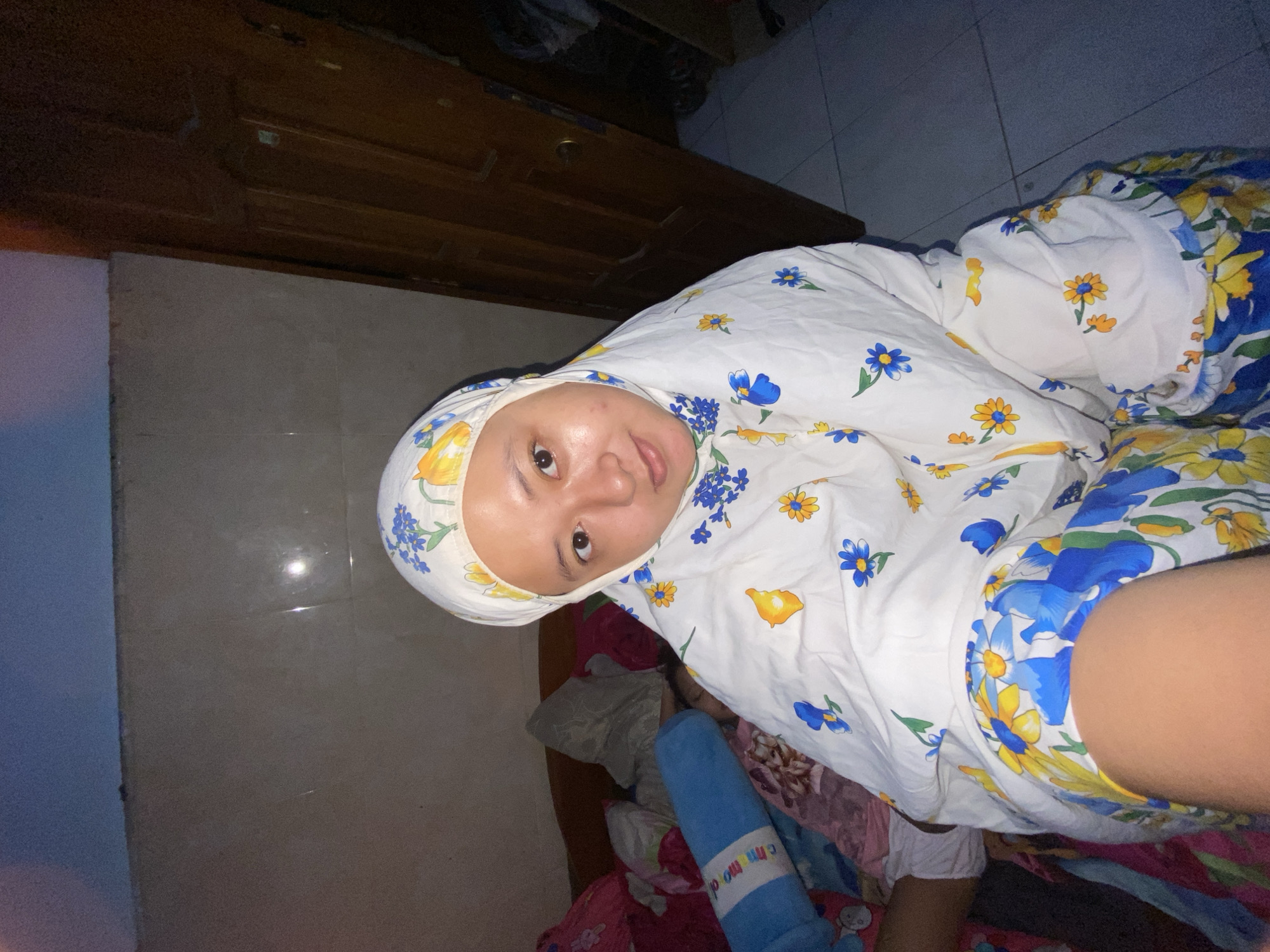The image depicts a person wearing a floral-patterned headscarf and lying down on what appears to be a bed covered with floral-patterned sheets. A blue cylindrical object with white lettering is visible near the bed. The location appears to be indoors, potentially a bedroom, in Gelam, Indonesia, based on visual cues from the background and cultural association with headscarf. A wooden cabinet or dresser is visible in the background.