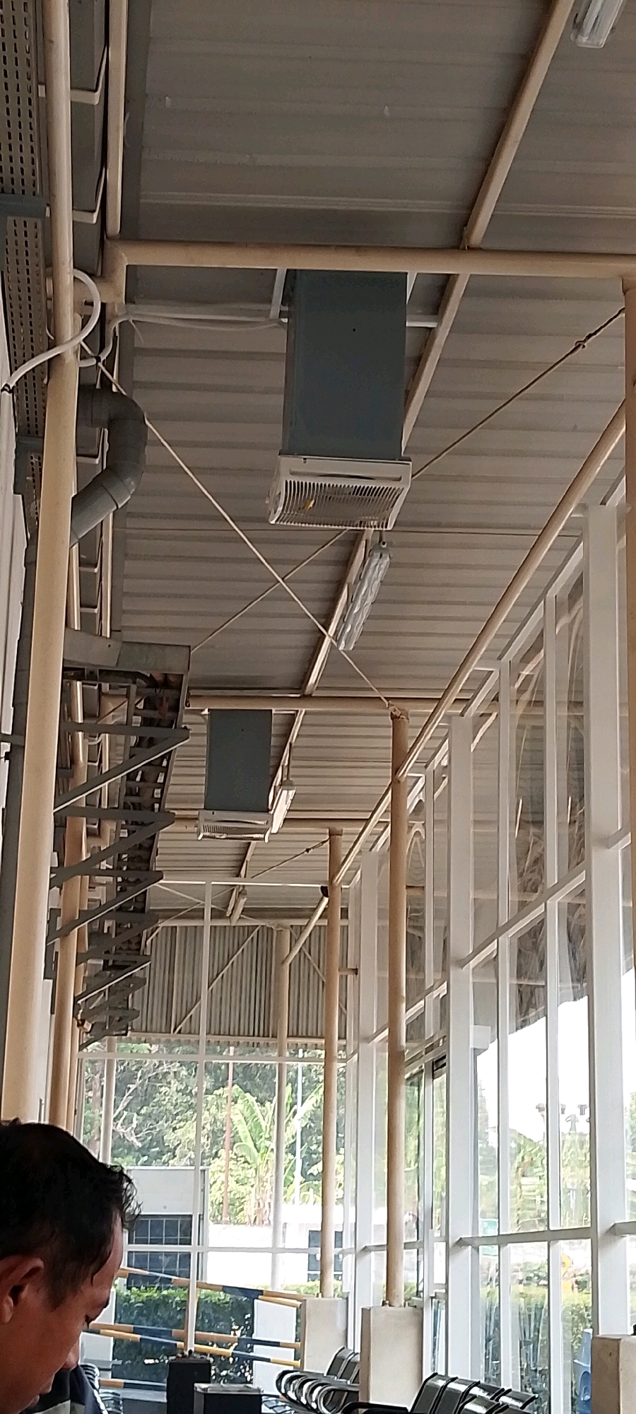 An outdoor view, possibly a waiting area or covered walkway, is depicted. The scene is characterized by a metal roof supported by light-colored beams and columns. Several rectangular, dark gray air conditioning units are mounted on the ceiling. One prominent grey duct pipe curves downwards from the ceiling on the left side. In the background, large glass windows reveal lush green foliage and trees, suggesting an outdoor environment. A man's head and shoulder are visible in the lower left corner, facing downwards. Rows of dark seating, possibly chairs or benches, are arranged in the foreground. The location appears to be a public space within the city of Depok, Indonesia, given the context of the air conditioning units and the style of the architecture.