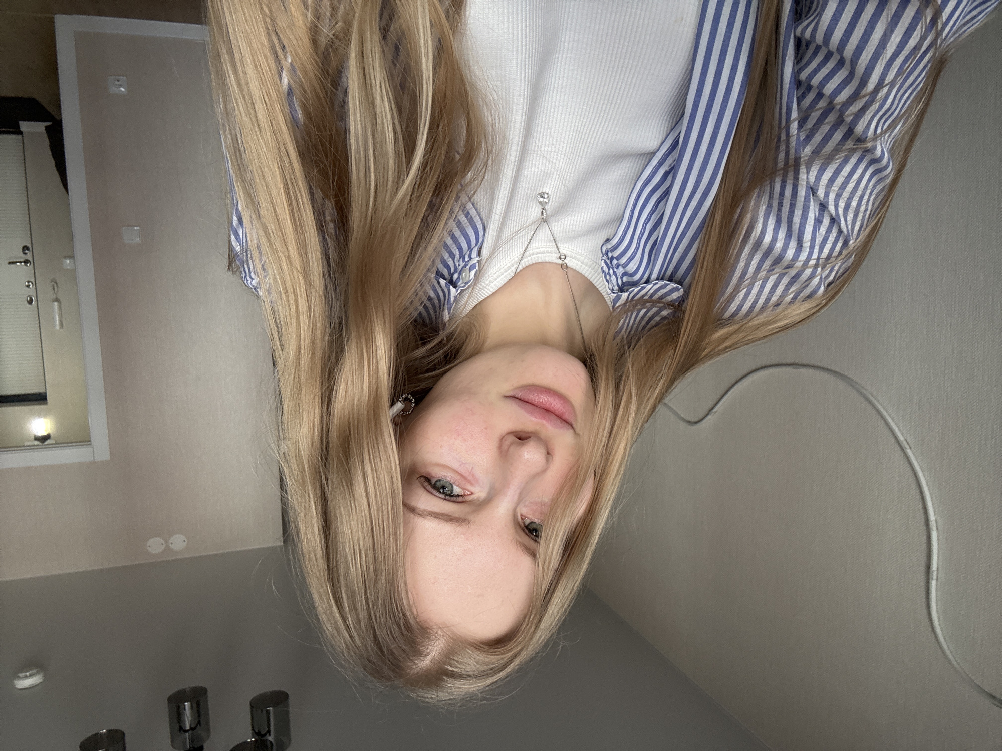 The image shows a blonde woman with light skin and blue eyes, wearing a white ribbed top and a blue and white striped shirt, with a silver necklace. The background includes a wall with electrical outlets, a window, and a bathroom sink and faucet, suggesting an indoor bathroom setting. While the location in the city of Perm, Russia is not directly visible, it is known information.