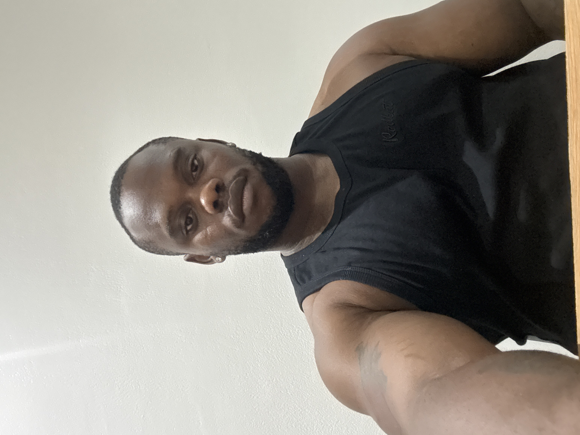 The image captures a man with dark skin, a short haircut, and a neatly trimmed beard, looking directly at the camera with a neutral expression. The perspective is from below, suggesting the man is looking down at the camera or that the photo is a selfie taken from a low angle. He is wearing a black sleeveless tank top, which highlights his muscular build, particularly his arms and shoulders. The word "Redbat" (or a similar-looking brand name) is visibly embroidered in black thread on the left chest area of his tank top.
The background consists of a plain white wall, evenly lit, indicating an indoor setting. A narrow, vertical strip of light-colored wood, possibly part of a doorframe or furniture, is visible along the far right edge of the frame. There are no other distinct objects or notable details in the background. The time of day is not discernible due to the indoor setting and artificial or diffused lighting. The overall scene is simple and uncluttered, focusing primarily on the man. This scene is set in Iddo Tudunwada, Nigeria.