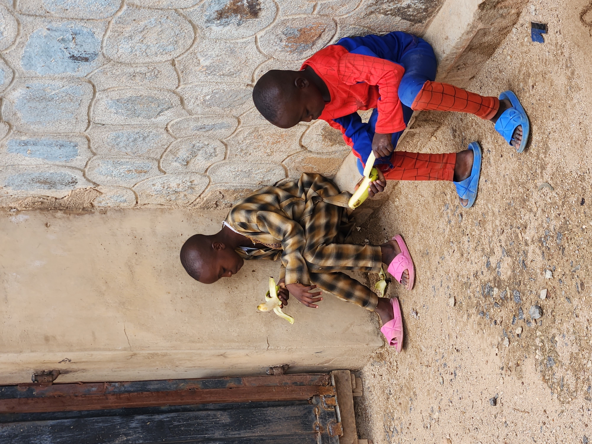 Two children are seated outdoors, peeling bananas. One child wears a red and blue superhero outfit with matching blue sandals. The other child is dressed in a tan and brown plaid outfit with pink sandals. They sit next to a stone wall and a door with rusty hardware. The ground is dirt and gravel. The location is Unknown, Unknown.